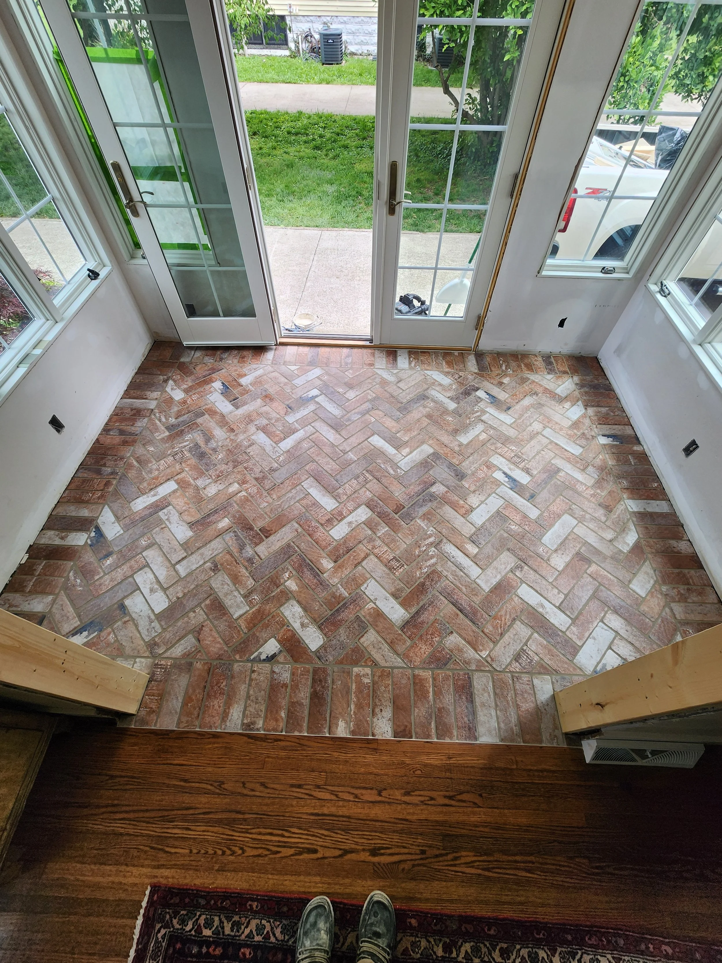 Inside view of a room with a brick floor, showing a glass door leading to a grassy outdoor area with trees and a sidewalk.
