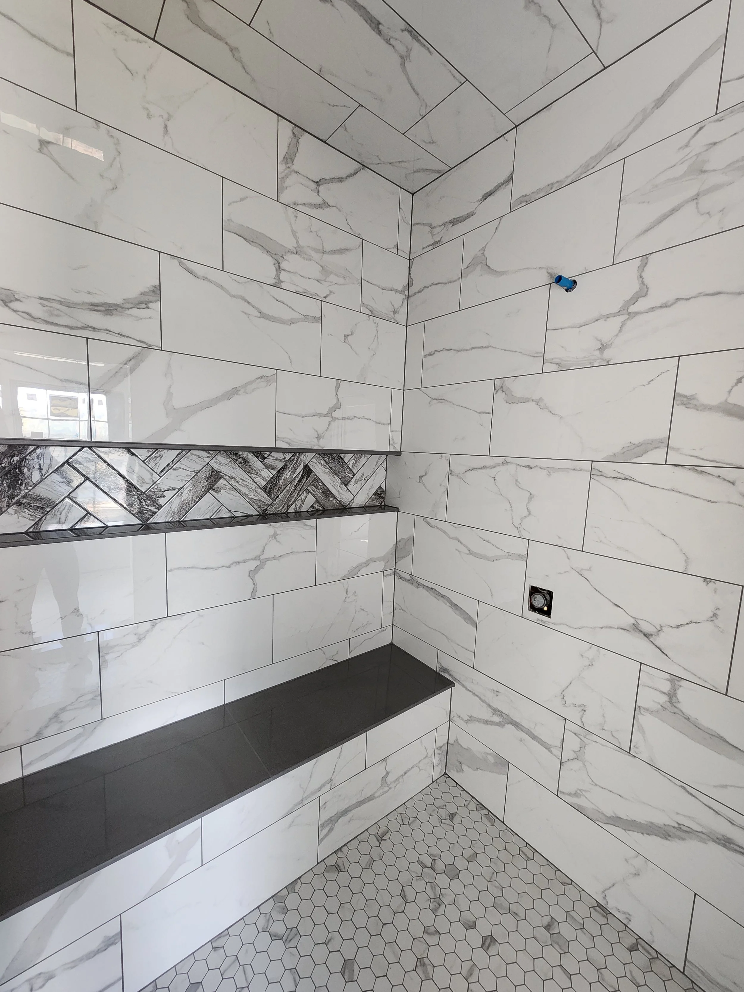A corner of a custom walk in bathroom with white marble-like tiles on the walls and gray tiles on the floor. There are built-in black shelves on the wall that creates an infinity niche, custom built bench and double shower heads