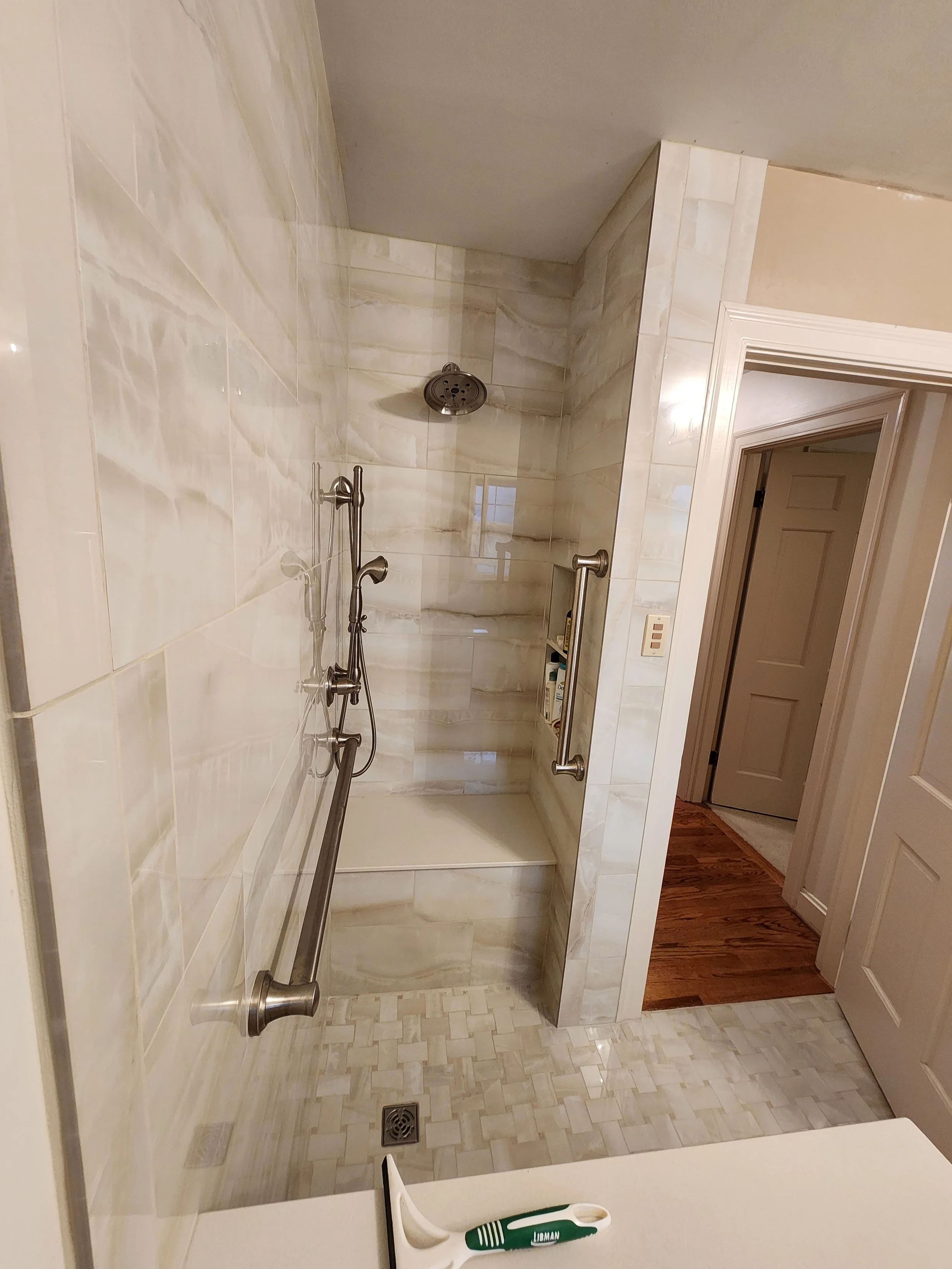 Bathroom with a walk-in shower featuring beige and white marble tiles, a grab bar, a showerhead, and a small built-in shelf holding various bottles. Shower is zero-entry and ADA accessible.