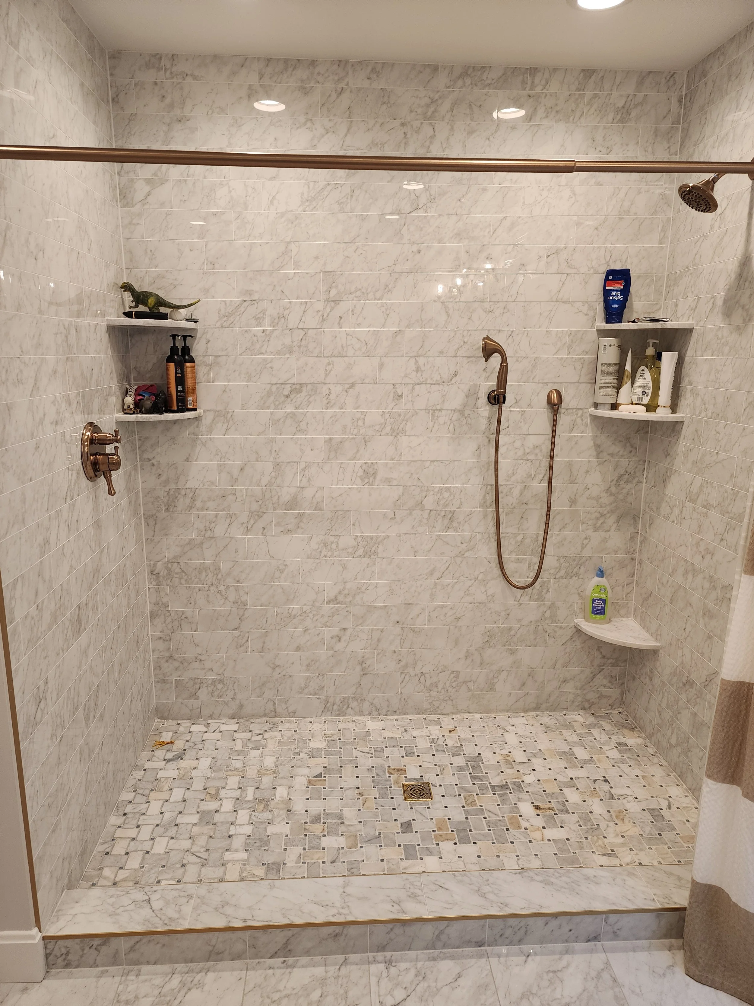 A walk-in shower with beige marble tiles on the walls and a mosaic tile floor. There are two shelves holding personal care products, a handheld shower head, and a ceiling-mounted shower head. A small drain is centered on the floor.