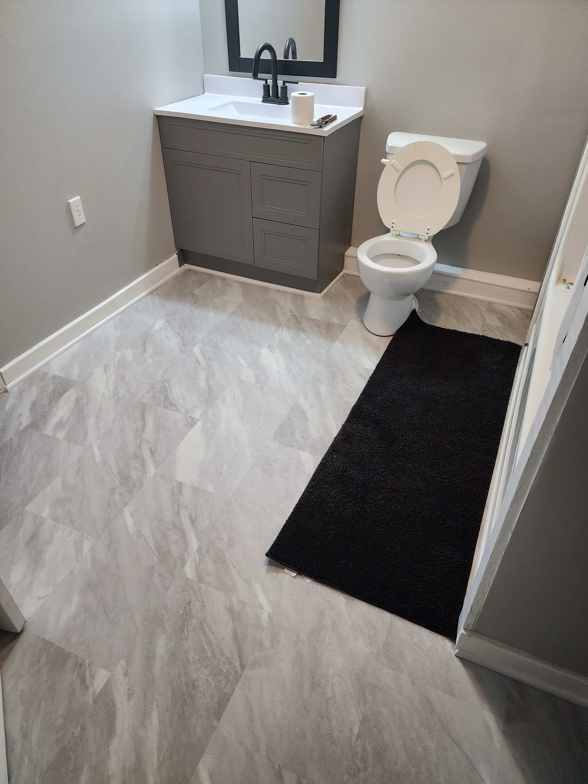 A bathroom with gray and white color scheme, featuring a toilet with an open lid, a gray vanity with a white counter, a black mirror above the vanity, and a black bath mat on a marble-like tiled floor. Personal items like a paper towel roll and a few