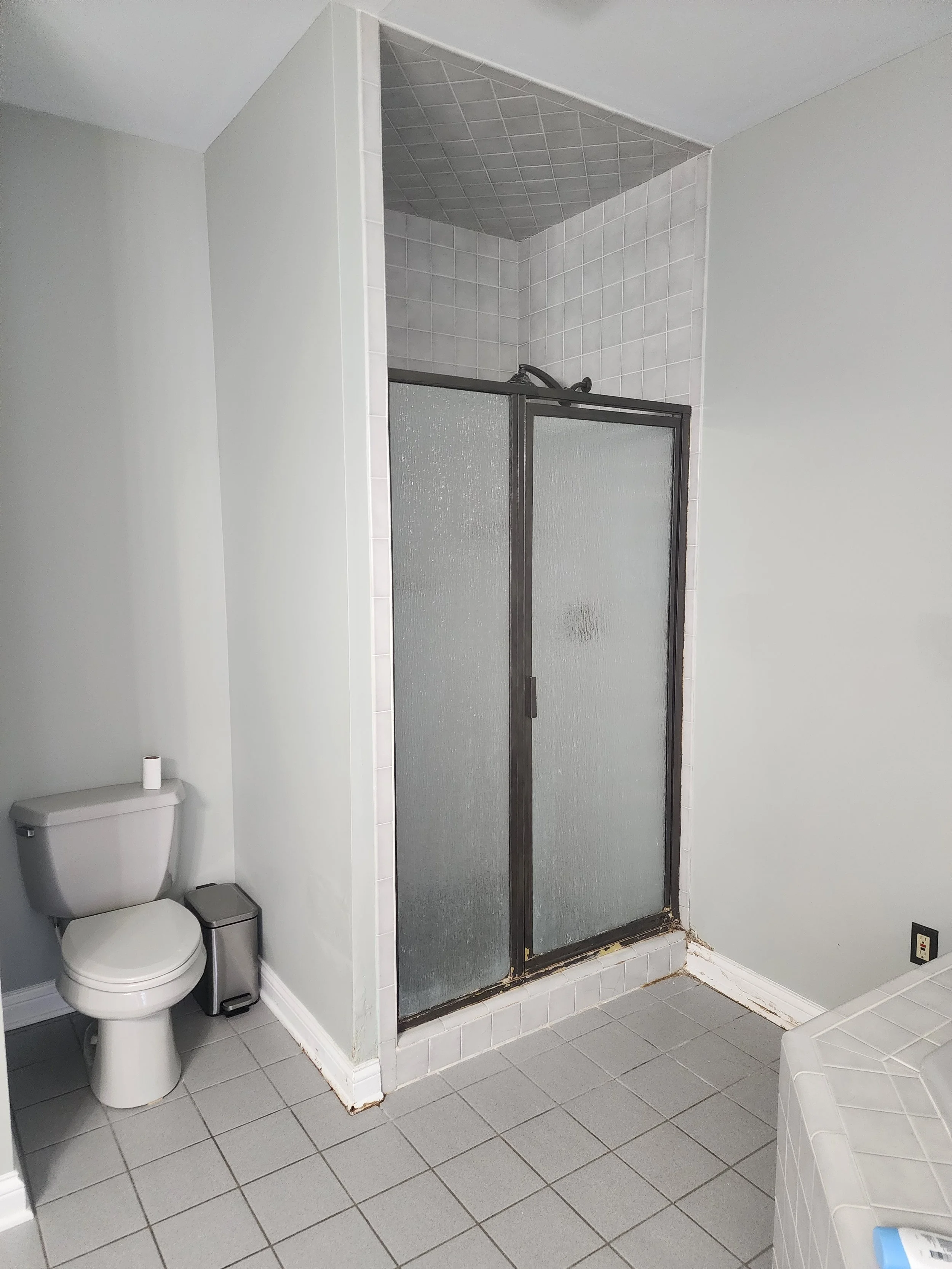A plain bathroom with a toilet on the left, a small trash can, a shower with textured glass doors, and gray tiled flooring and walls.