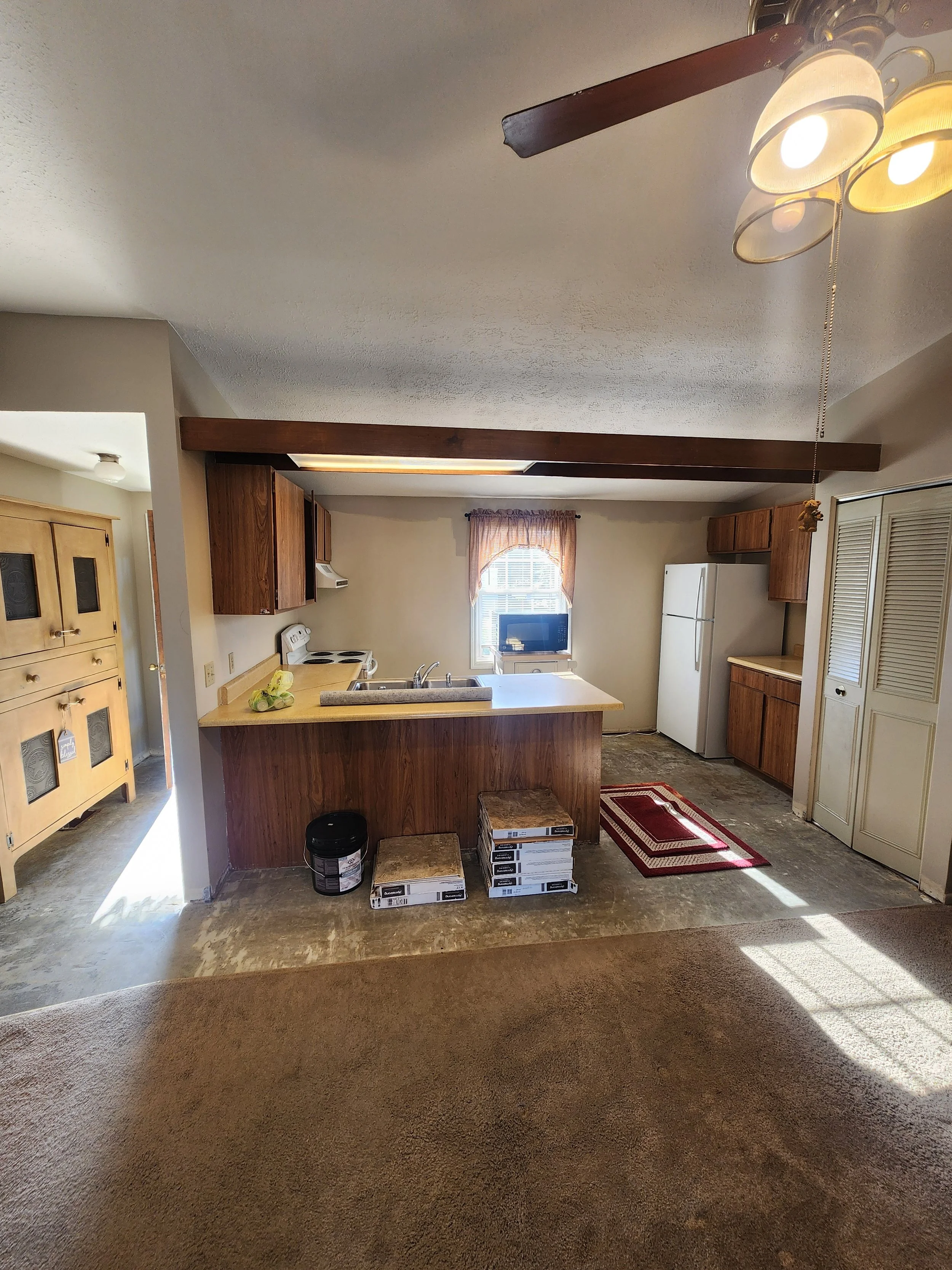 View of a kitchen with wooden cabinets, a white refrigerator, a small window, and an island with a sink. There are boxes on the floor, a small red rug, and a beige carpet in the foreground. Sunshine streams in from the right.