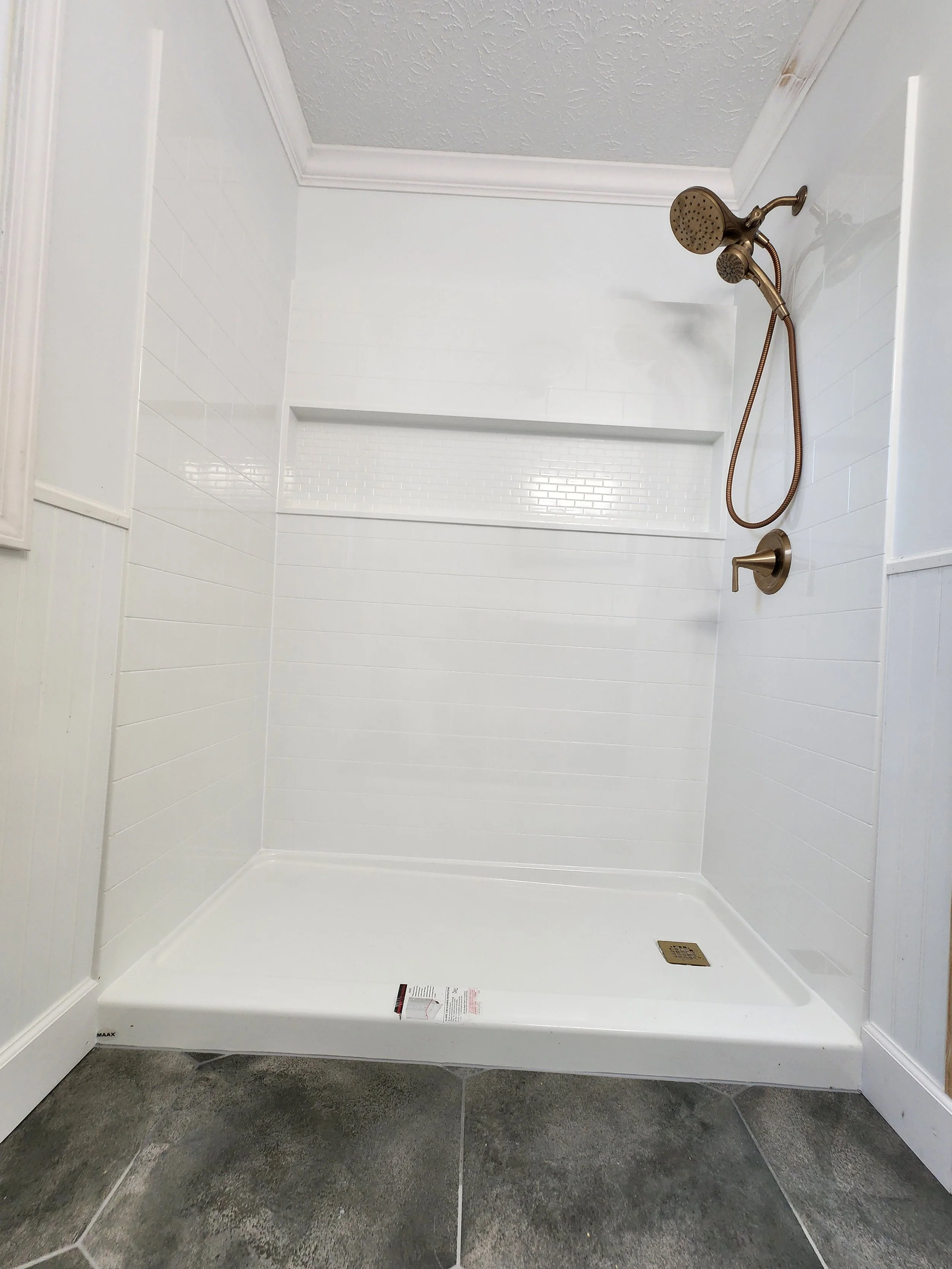 Prefabricated, low cost, walk-in shower with faux white tiled walls, a handheld showerhead, and a drain on gray tiled floor.