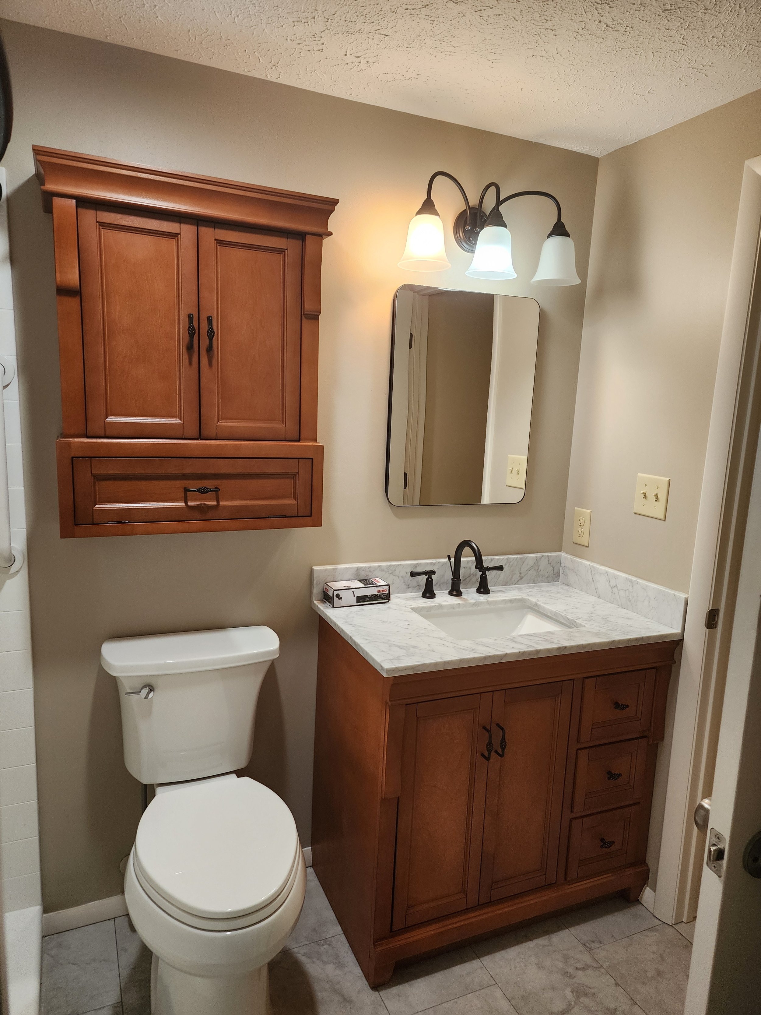 A bathroom with a toilet, wooden cabinet, marble countertop sink, mirror, wall-mounted light fixture with three shades, and electrical outlets.
