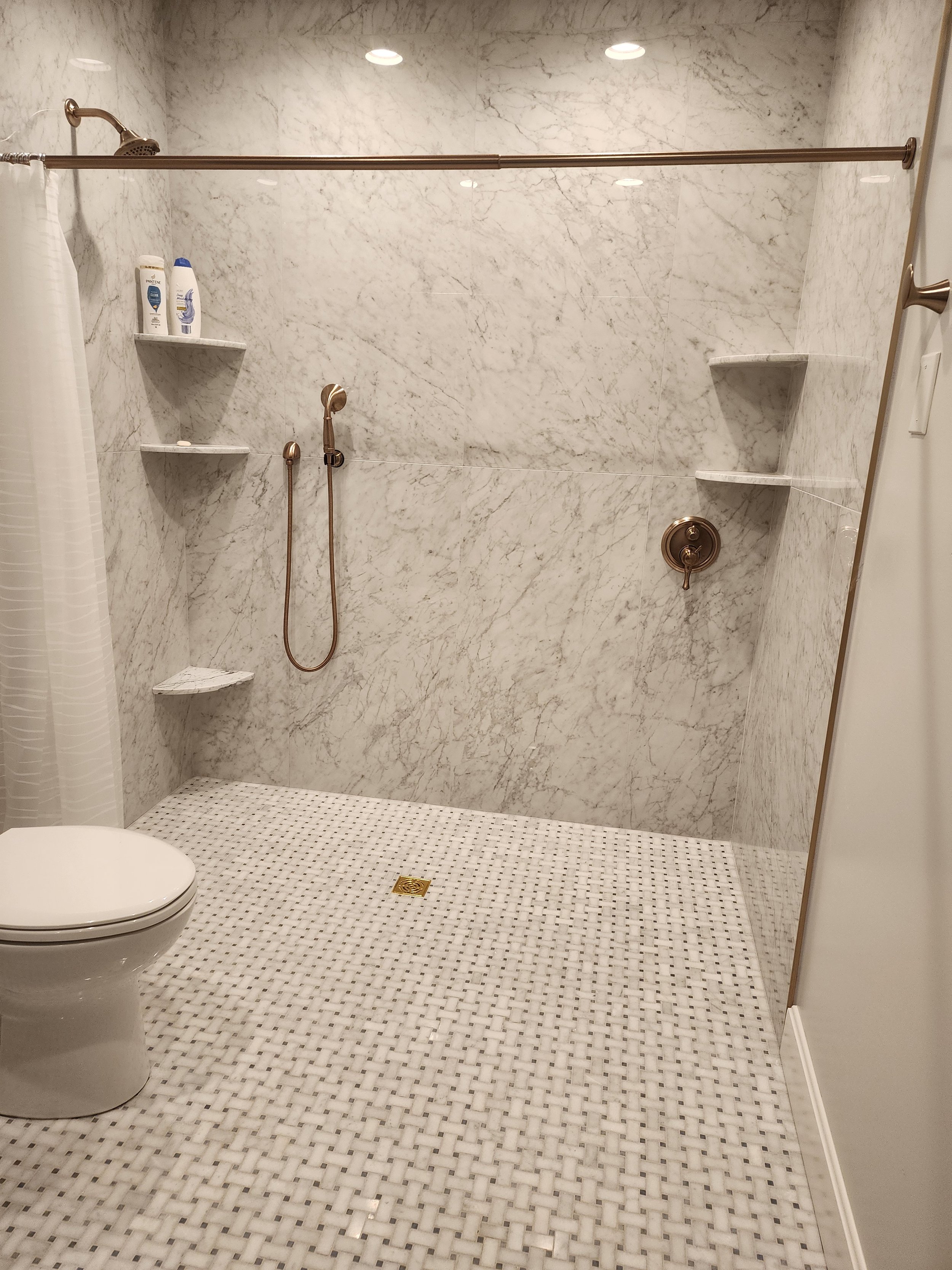 A zero entry, ada accessible wet room shower area with marble walls, a showerhead, two small shelves with toiletries, a handheld shower hose, a brass faucet, a drain on a white tiled floor, and a partial view of a toilet and a shower curtain.