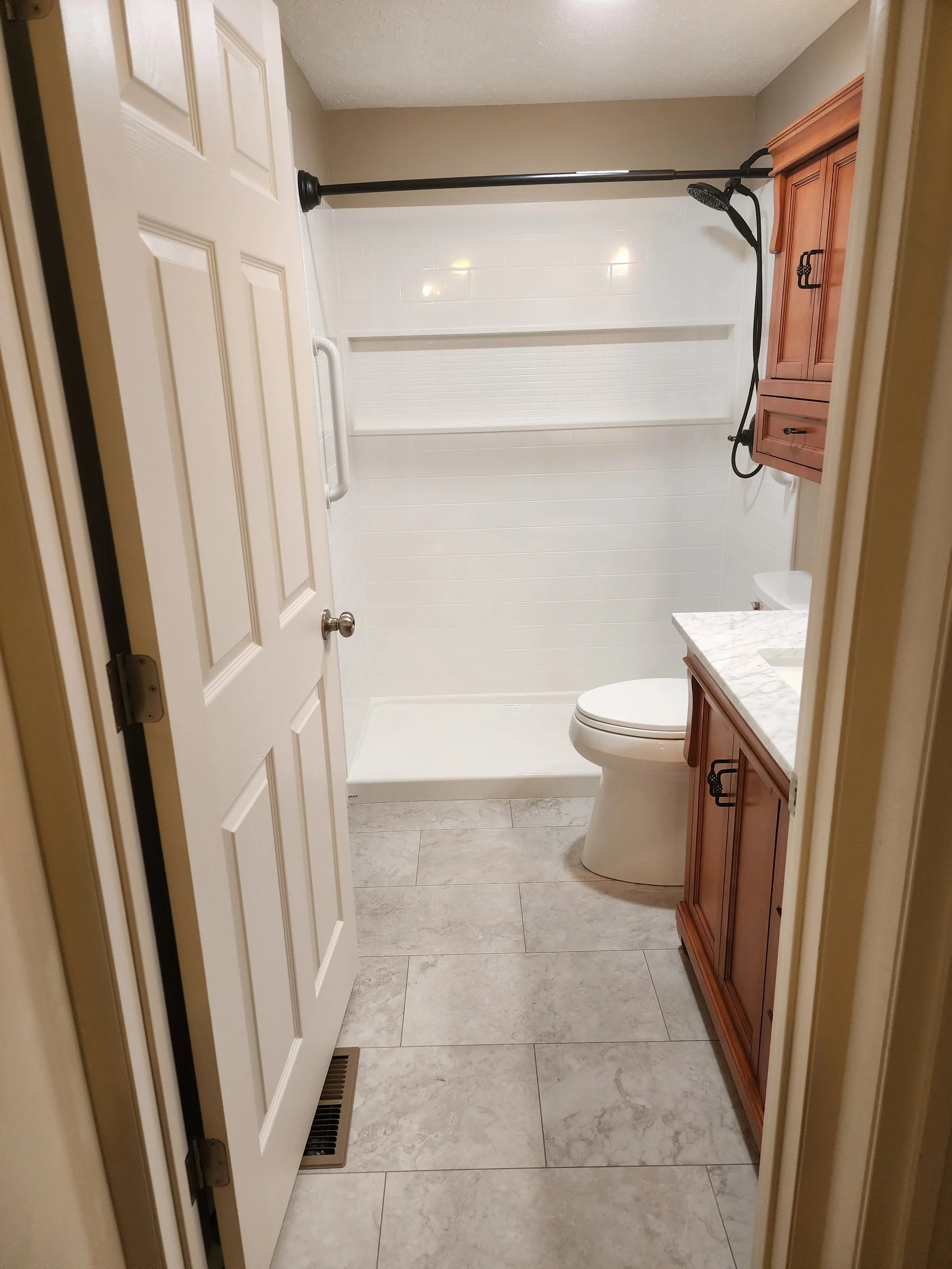 A clean bathroom with a white door partially open, a marble countertop with a sink, a wooden cabinet, a toilet, and a tiled shower area with a black showerhead and a black shower curtain rod.