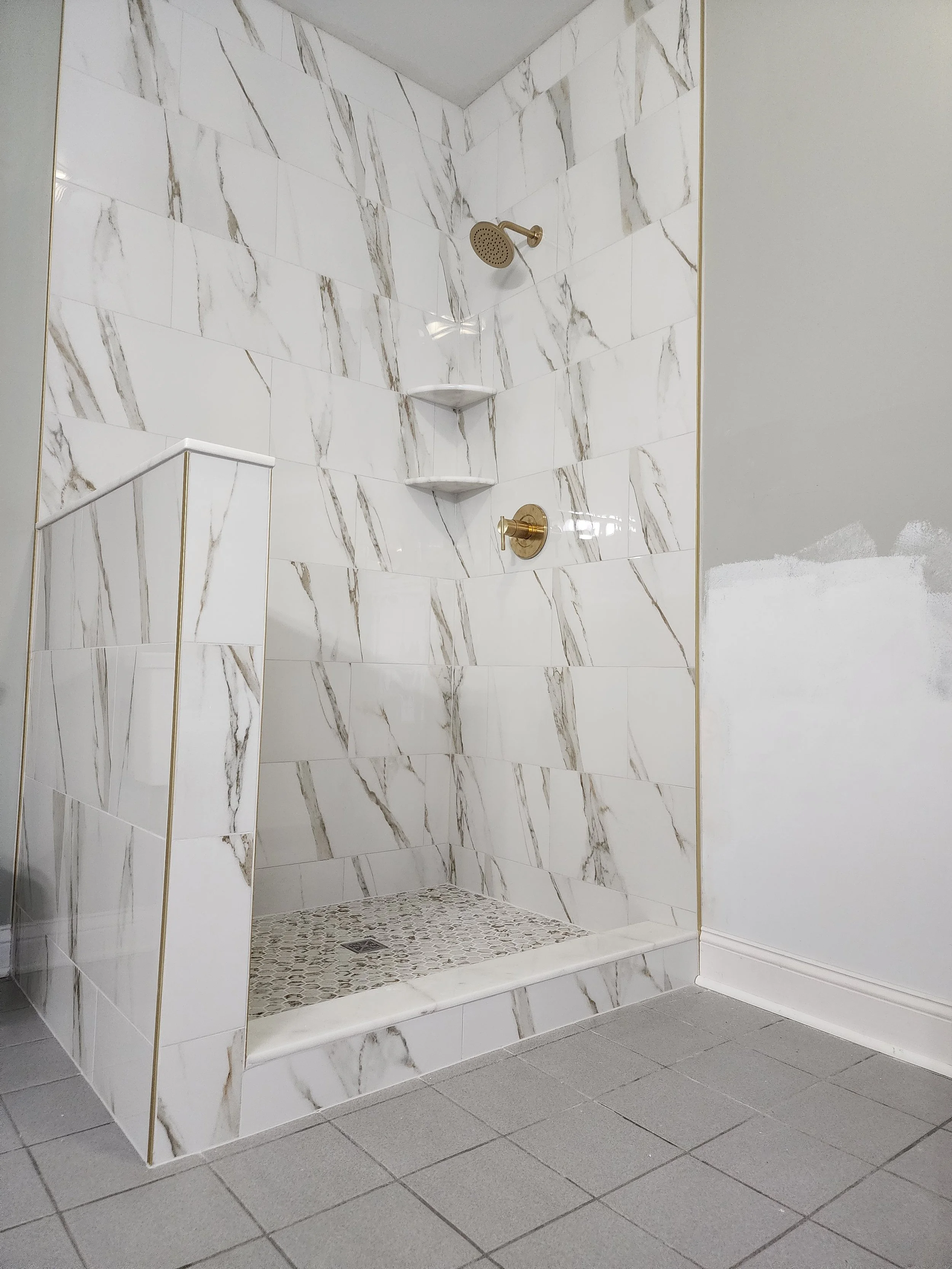 A walk-in shower with white marble tiles that have gray and gold veining, featuring two built-in white corner shelves, a gold showerhead, and a gold faucet.