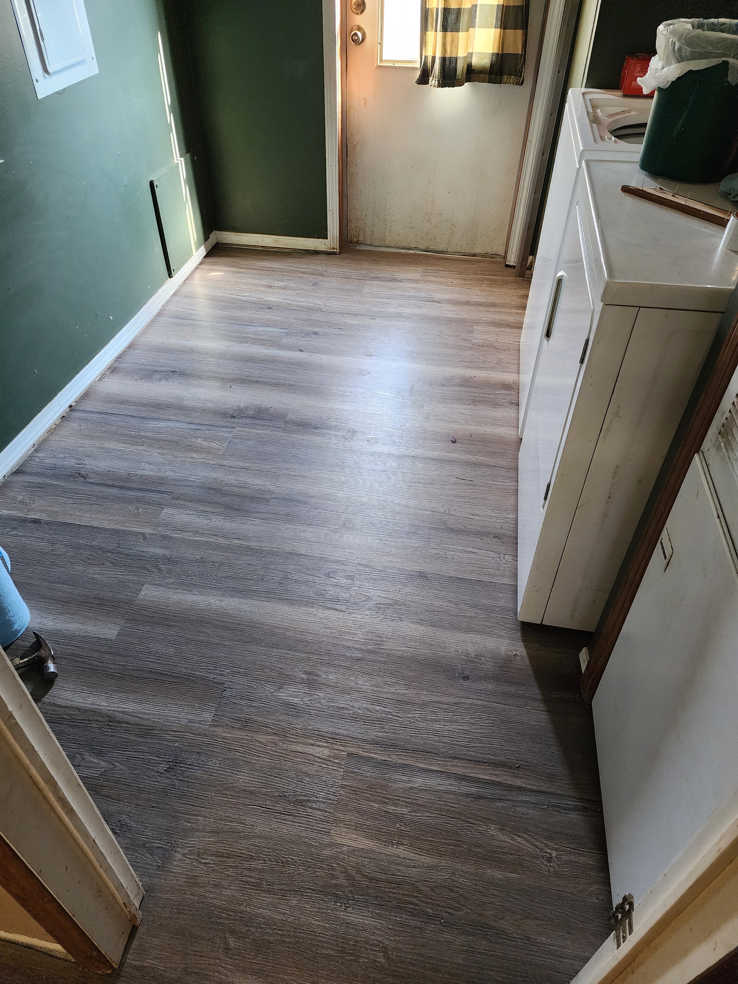 Empty room with wood laminate flooring, green and beige walls, a small window with plaid curtains, and laundry appliances on the right side.