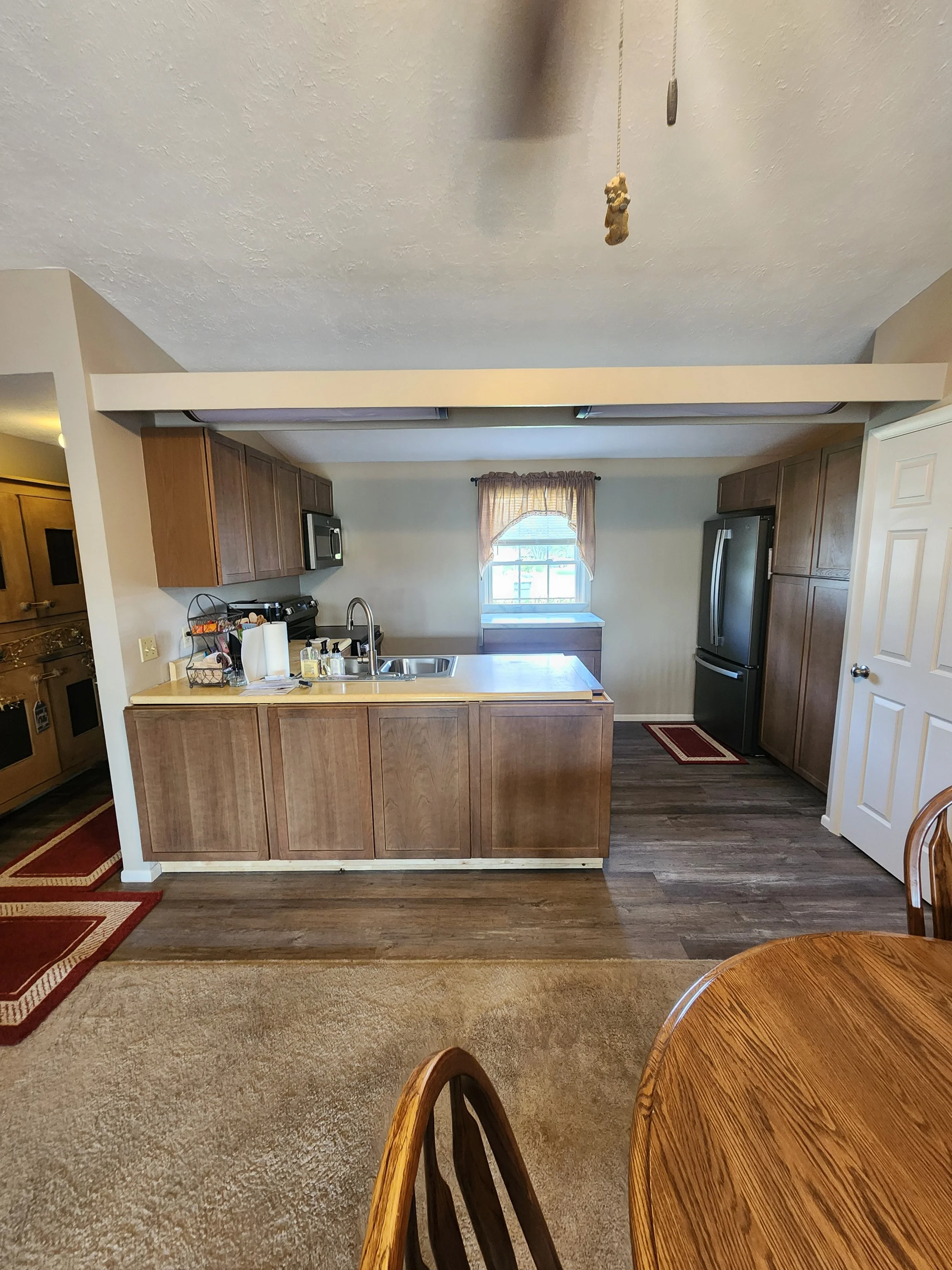 A view of a kitchen with wooden cabinets, black appliances, a window with a curtain, and a kitchen island with a faucet. Part of a dining table and chairs are also visible in the foreground.