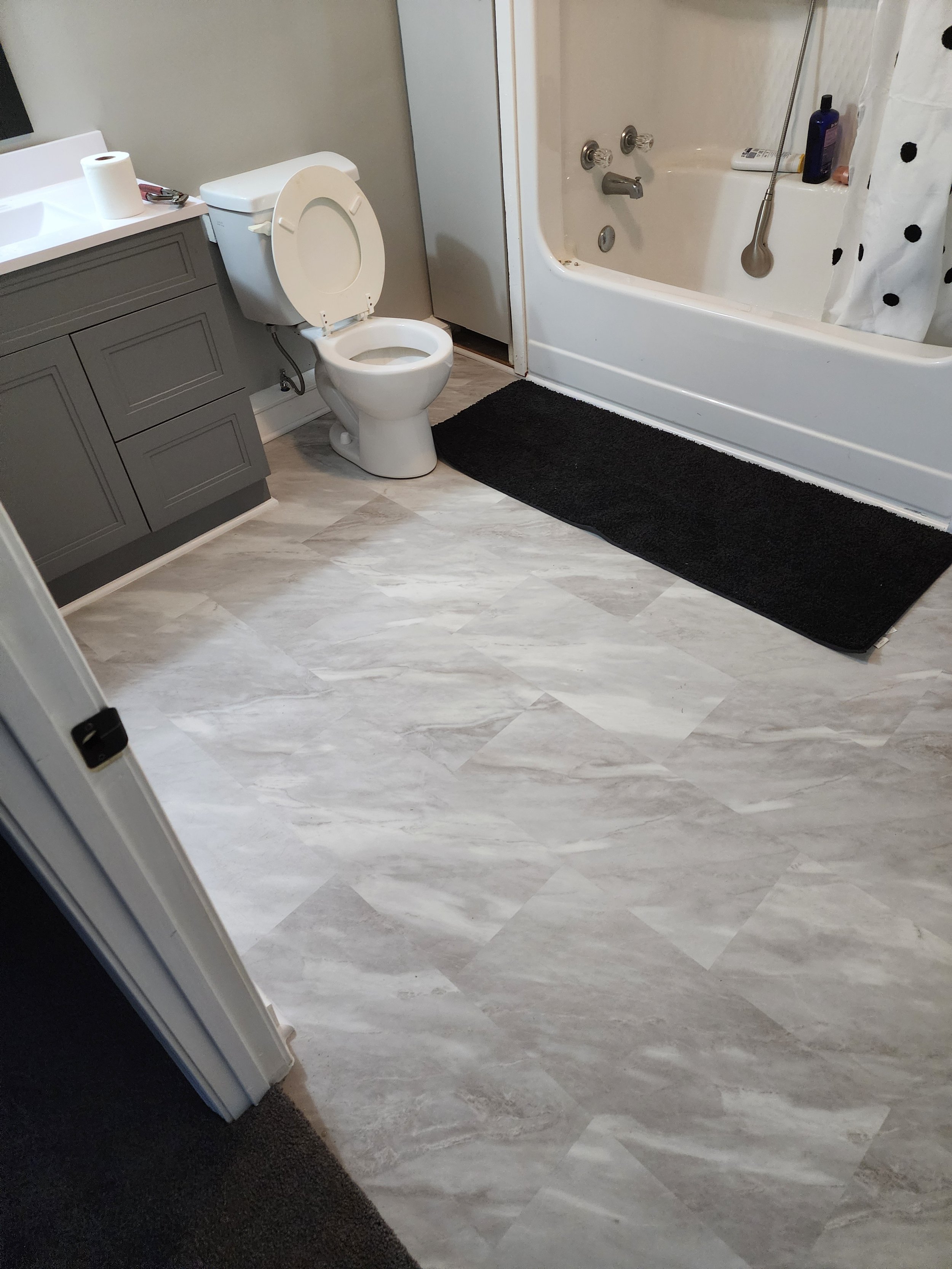 A bathroom with a toilet, a bathroom vanity, and a bathtub. The floor has light-colored tiles, and there is a black rug in front of the bathtub.