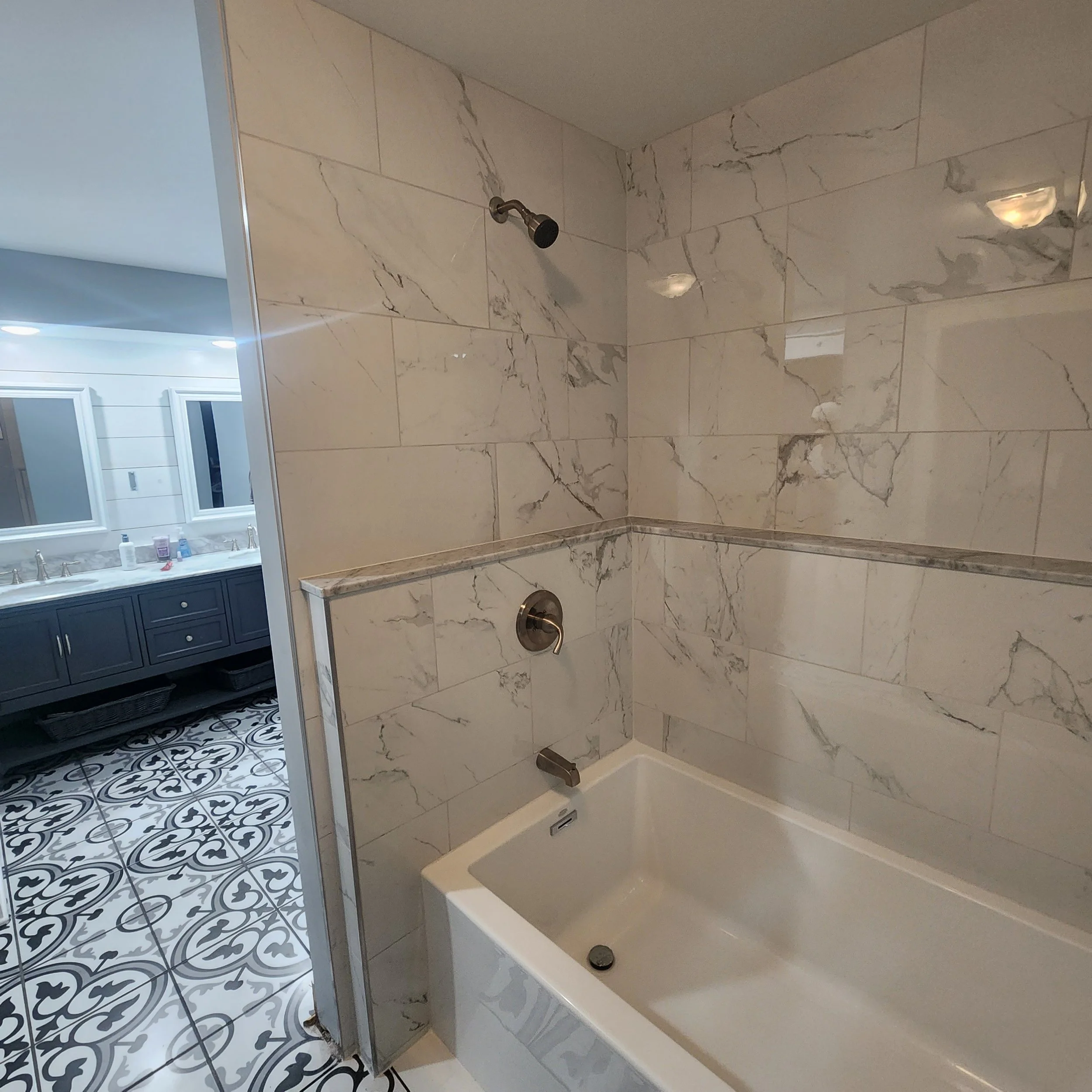 Bathroom with marble tiled walls, a recessed bathtub, and an adjoining shower area with a showerhead on the wall. The bathroom features contrasting patterned and solid-colored floor and wall tiles.