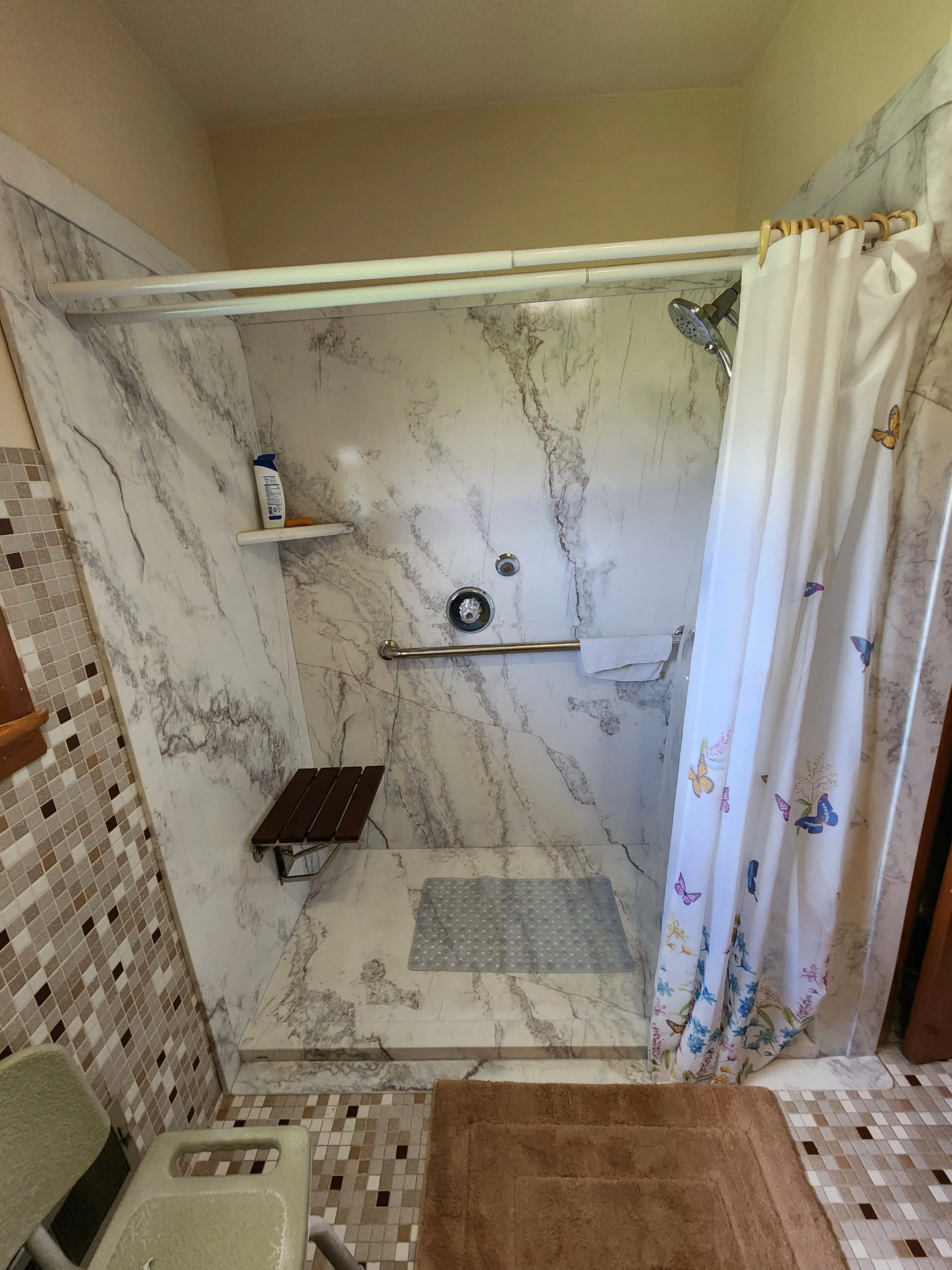 A prefabricated, zero entry, ada accessible bathroom shower area with marble walls, a white shower curtain with butterfly patterns, and a brown wooden bench. , a towel hanging on a rail, a gray bath mat, and mosaic tile accents on the floor and wall.
