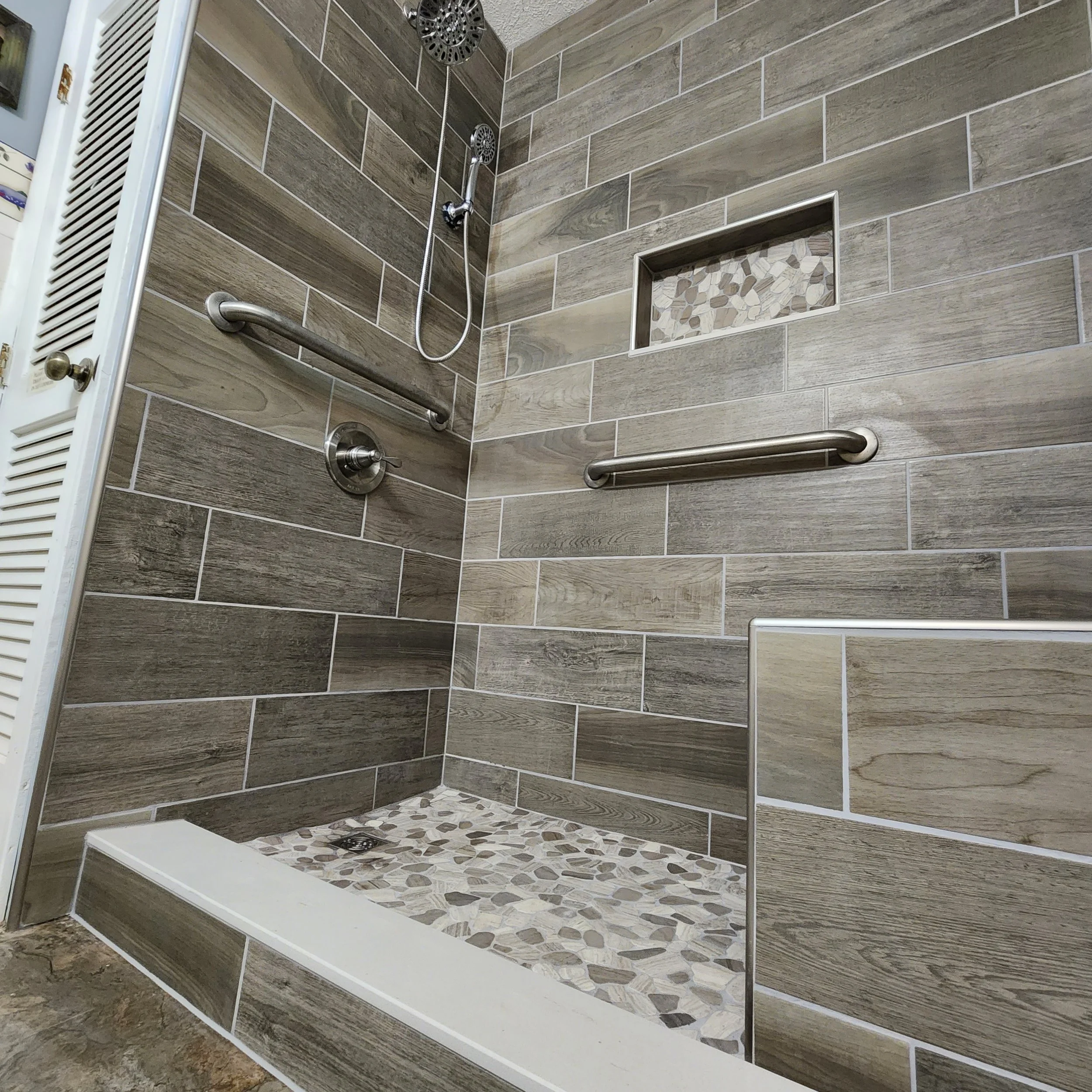 Shower with beige and gray wood-like tiles, pebble mosaic floor, wall niche with pebble design, and stainless steel grab bars.