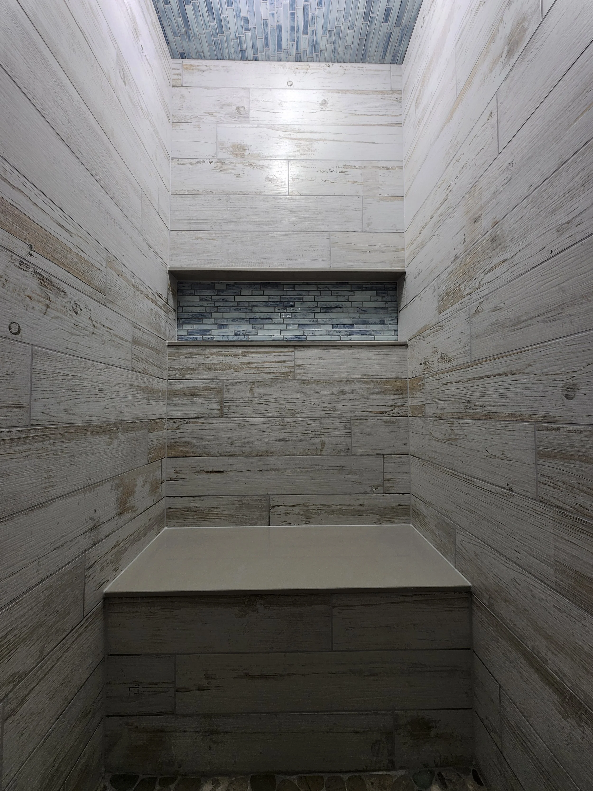 Interior view of a shower stall with light-colored wood-look tile walls, a ledge with dark gray subway tiles, and a pebble stone base.