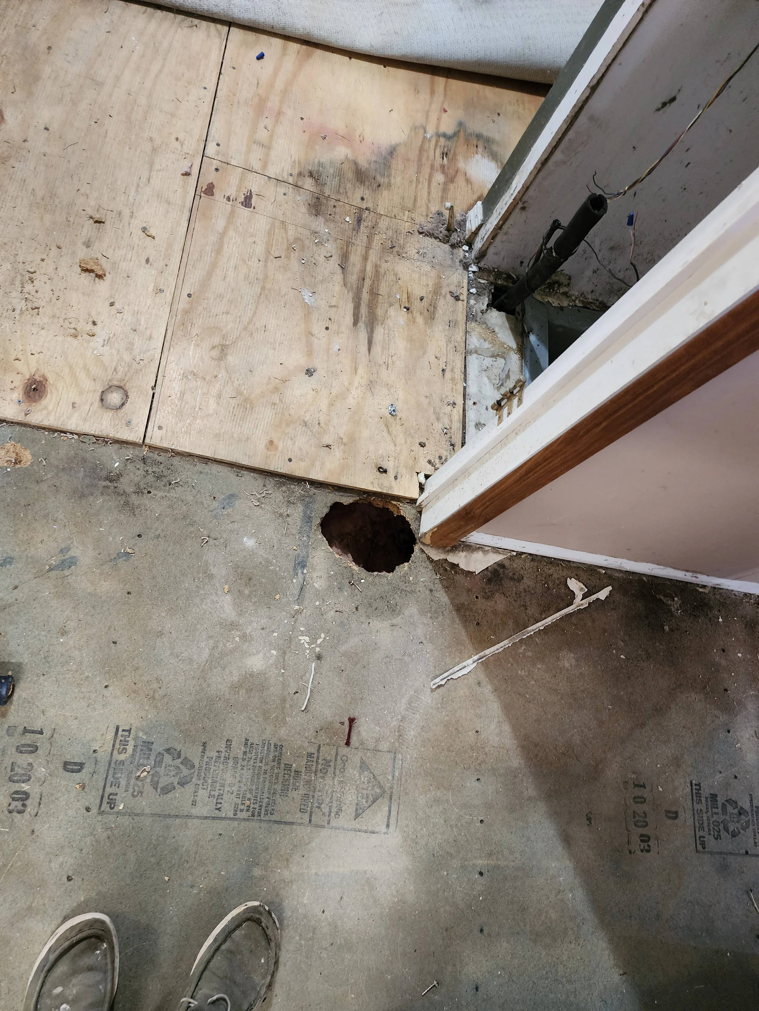 Construction site with plywood flooring, a hole in the concrete floor, and a partially built wall with exposed wiring and a mounted pipe or conduit.