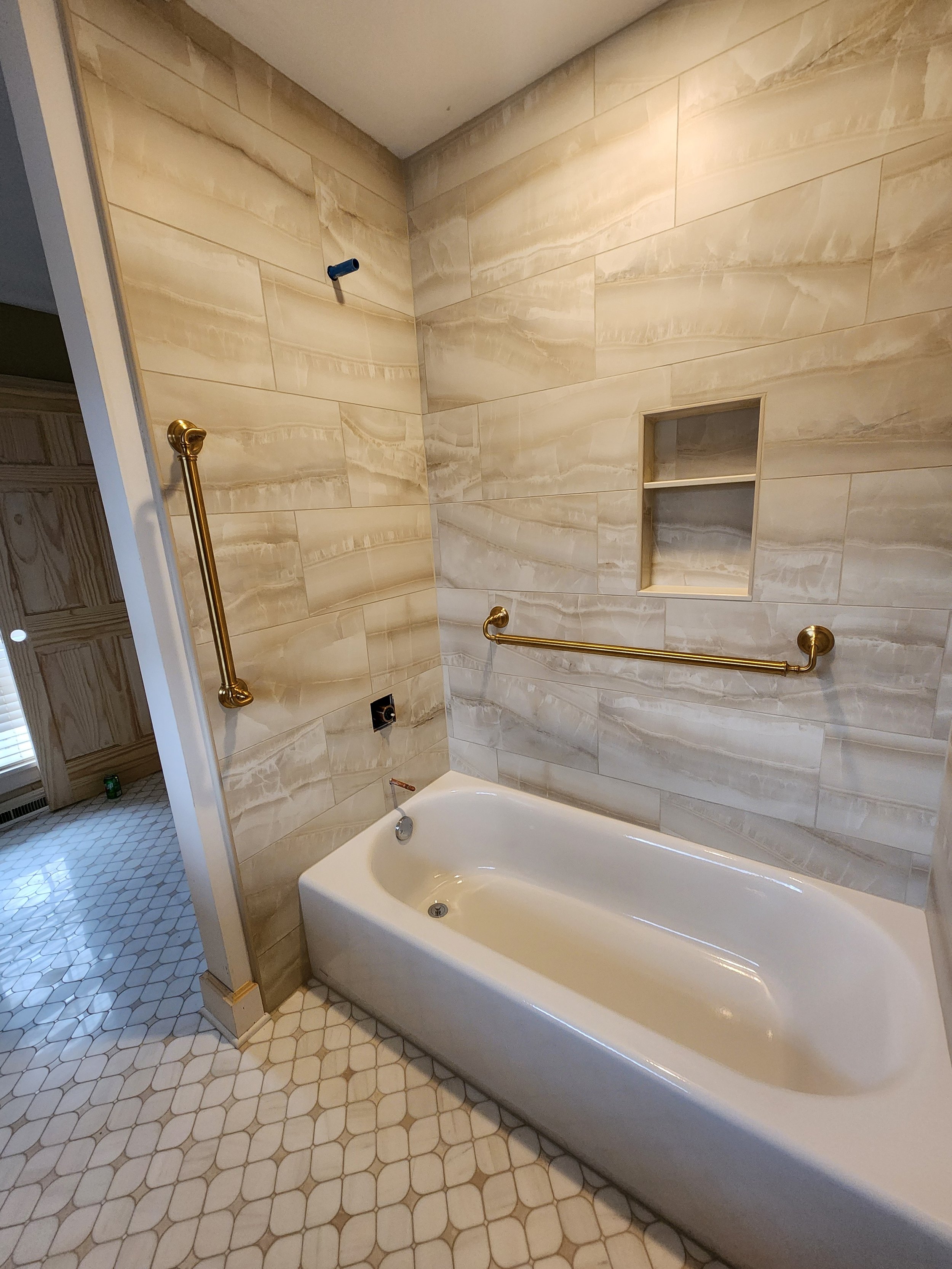 Bathroom with a corner bathtub, marble tile walls, a built-in shelf, and brass grab bars. The floor has white hexagonal tiles, and there is a small window to the left.