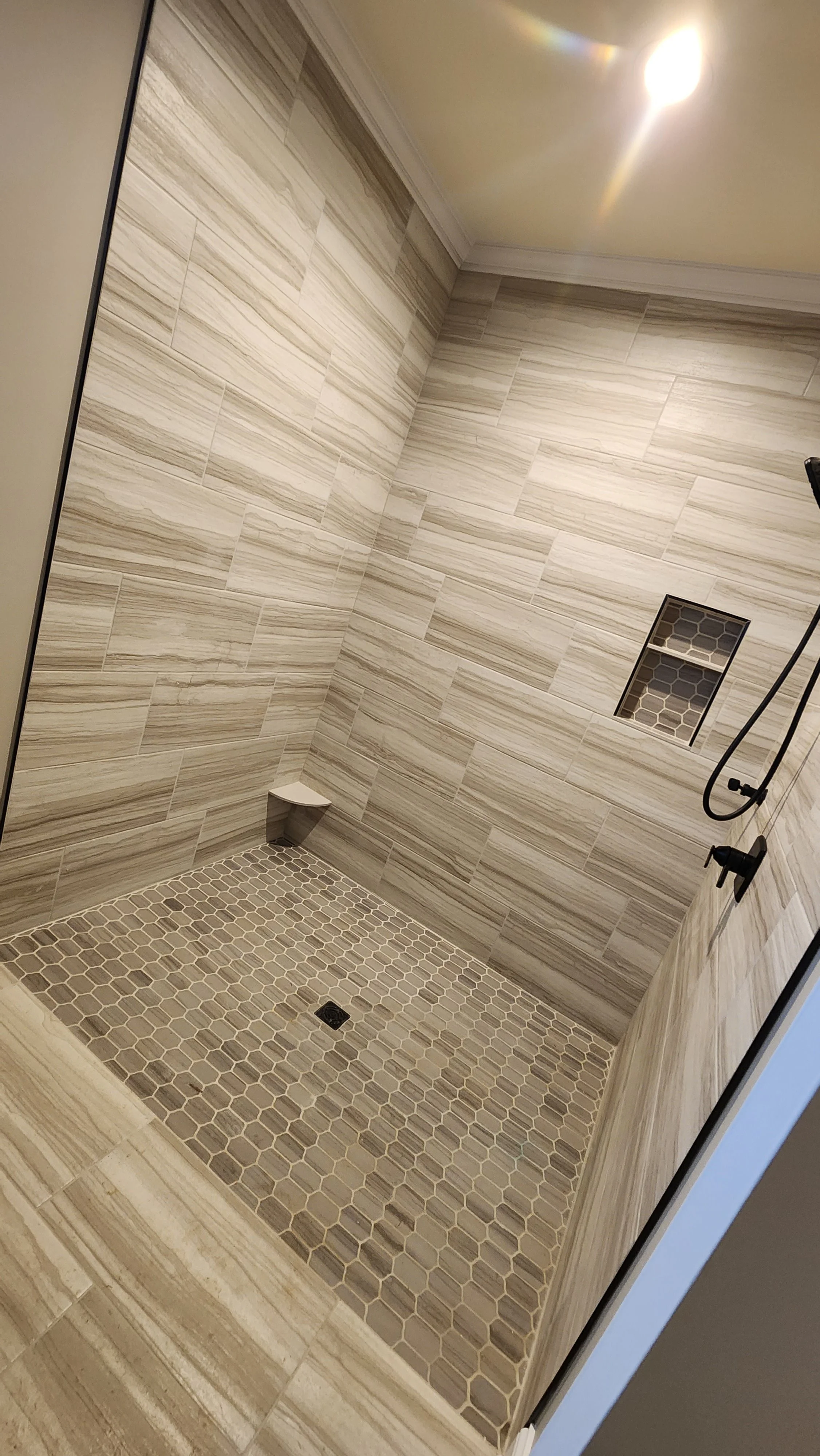 A modern shower with beige and cream porcelain striped tiles on the walls, a small corner shelf, a built-in niche with a honeycomb pattern, a black showerhead and controls, a drain, and a hexagonal tile floor.