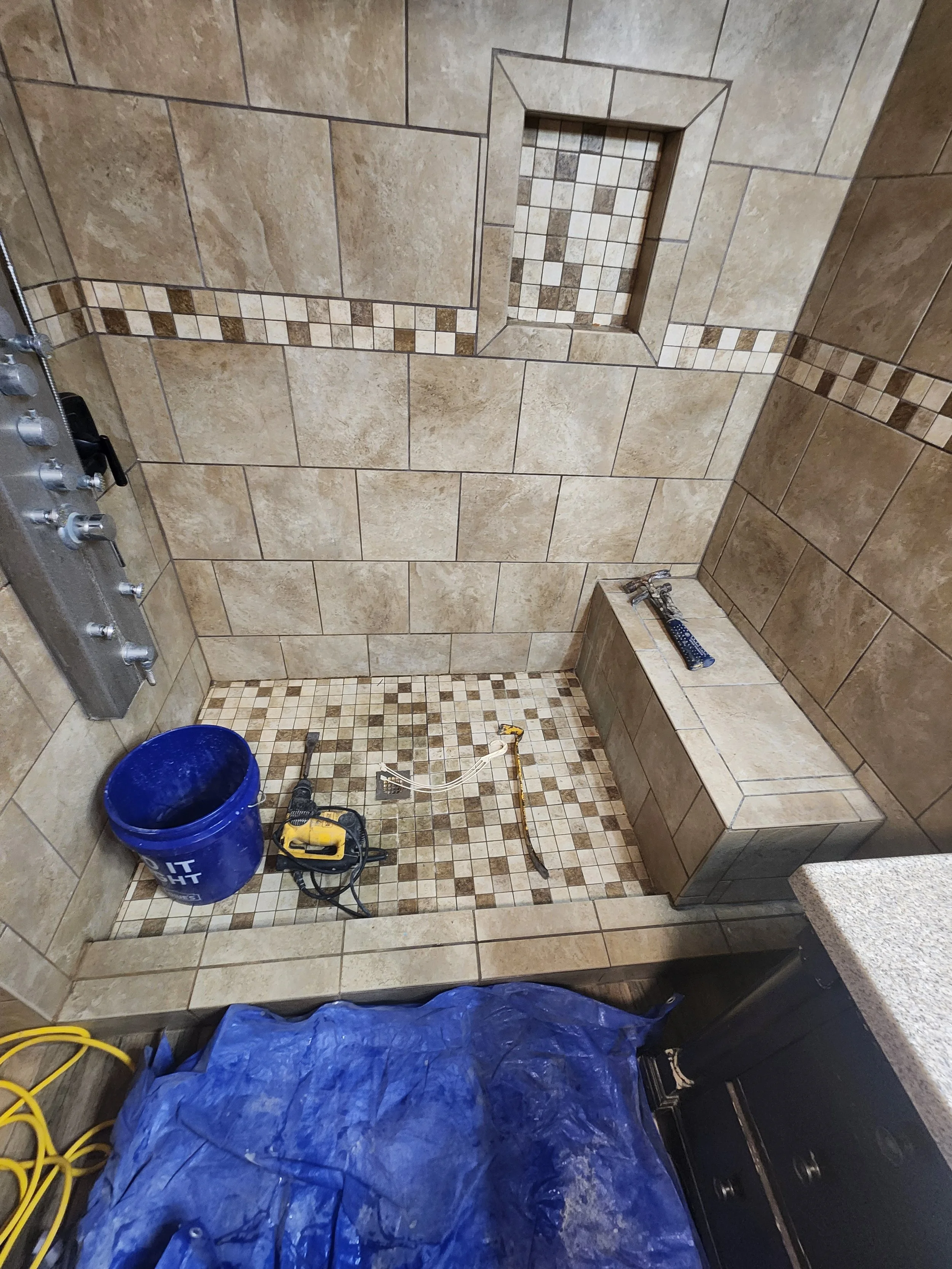A partially completed tiled shower with beige and brown tiles, a built-in niche, and construction tools including a blue tarp, yellow extension cord, and drill.
