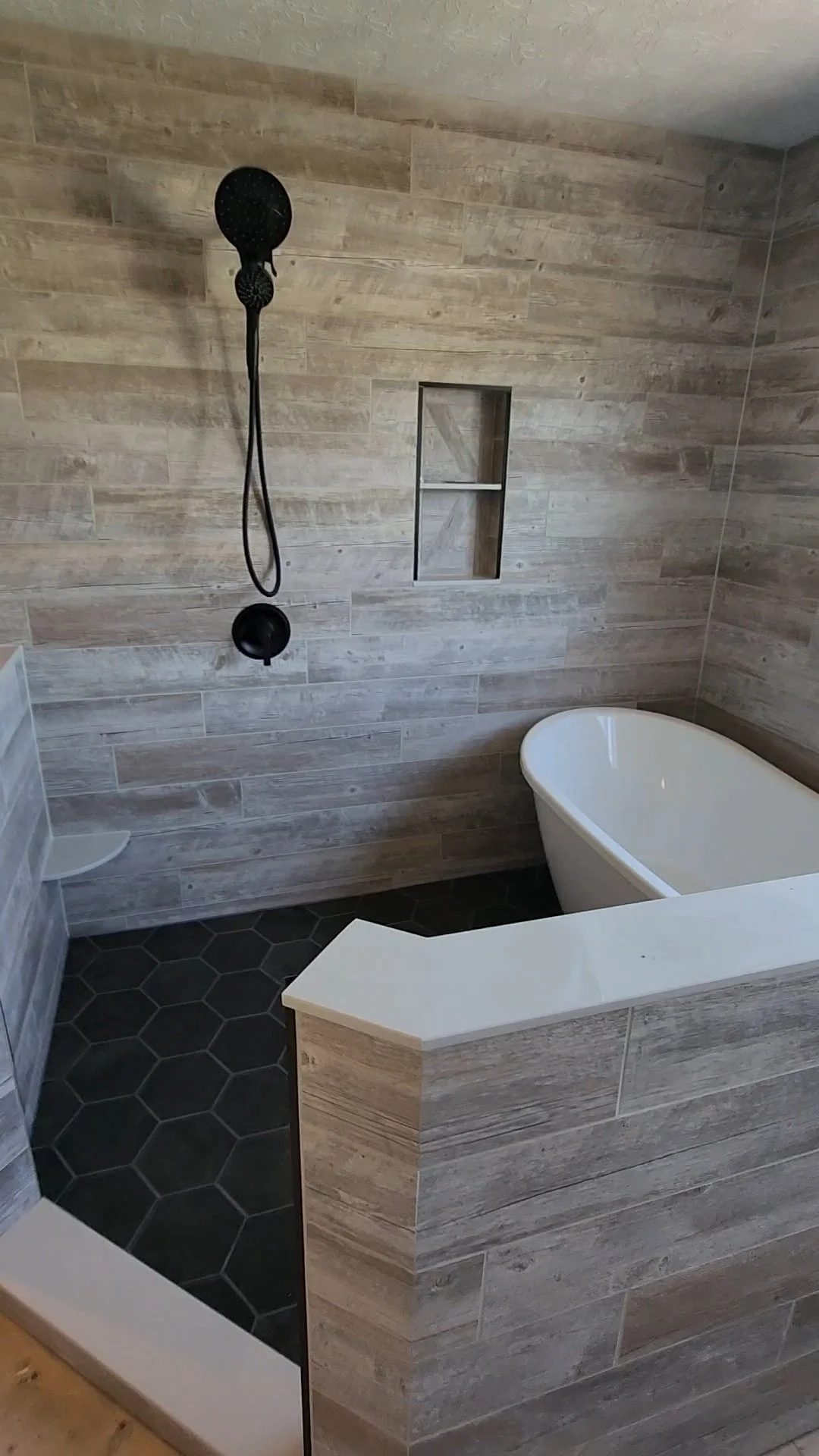 Modern bathroom with a walk-in shower featuring black fixtures, a small built-in shelf, and a freestanding bathtub with wood-like tile walls and hexagonal black floor tiles.