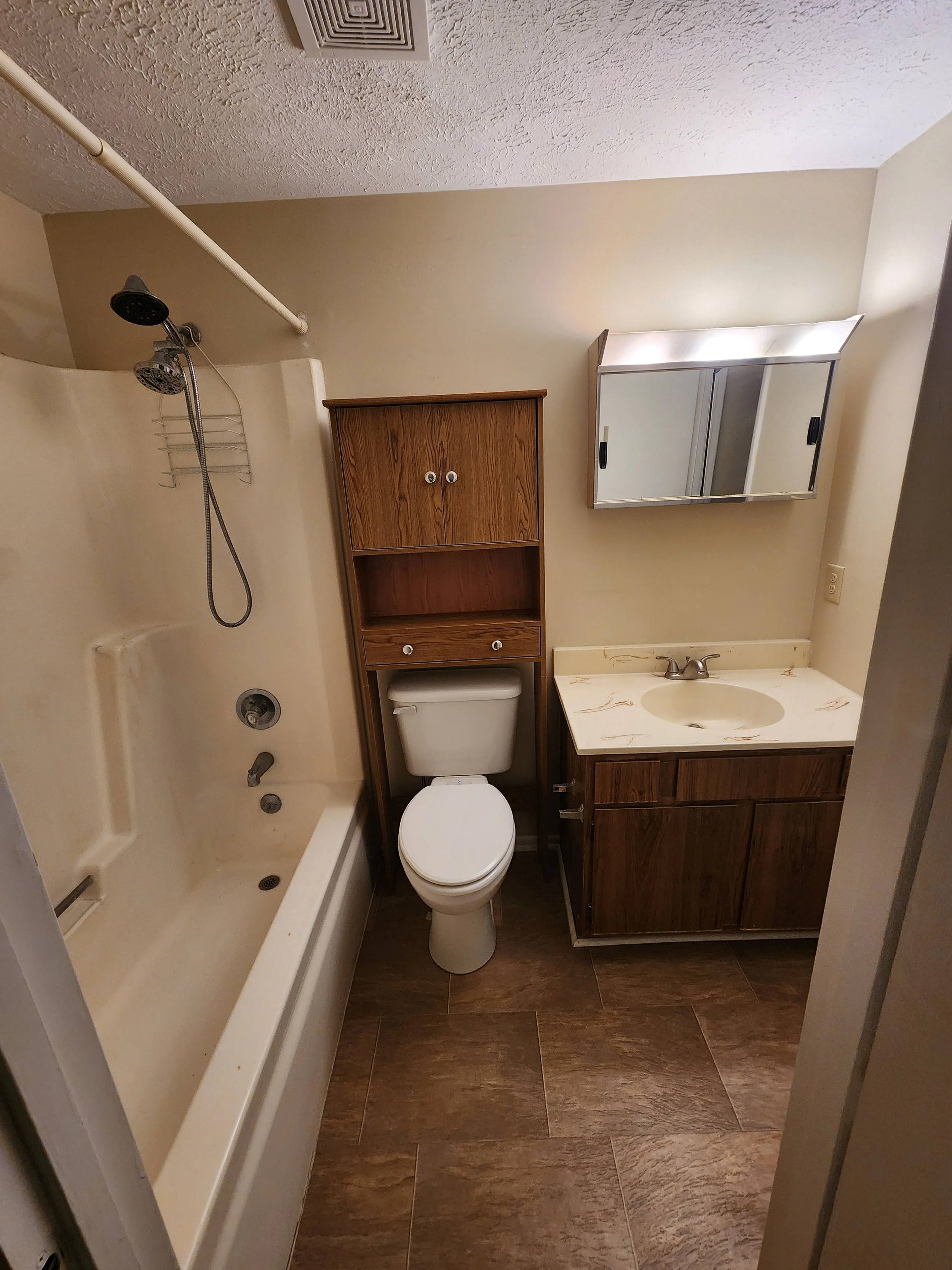 A small bathroom with a bathtub, showerhead, toilet, wooden cabinet, mirror, and a beige countertop with a sink, beige walls, and brown wooden floor tiles.