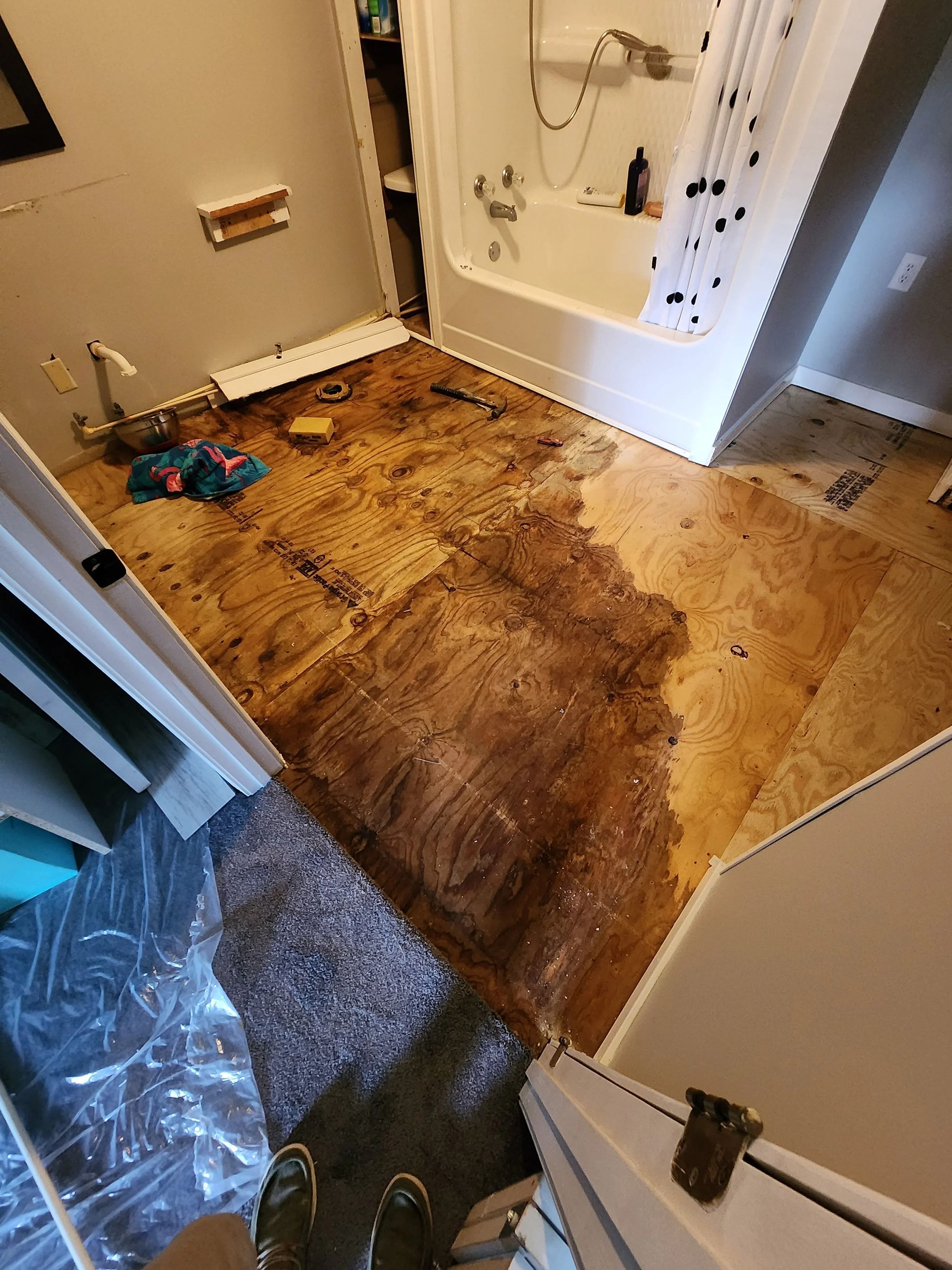 Bathroom under renovation with removed flooring, exposing subfloor, a bathtub with a shower curtain, tools on the floor, and a person standing in front taking a photo.