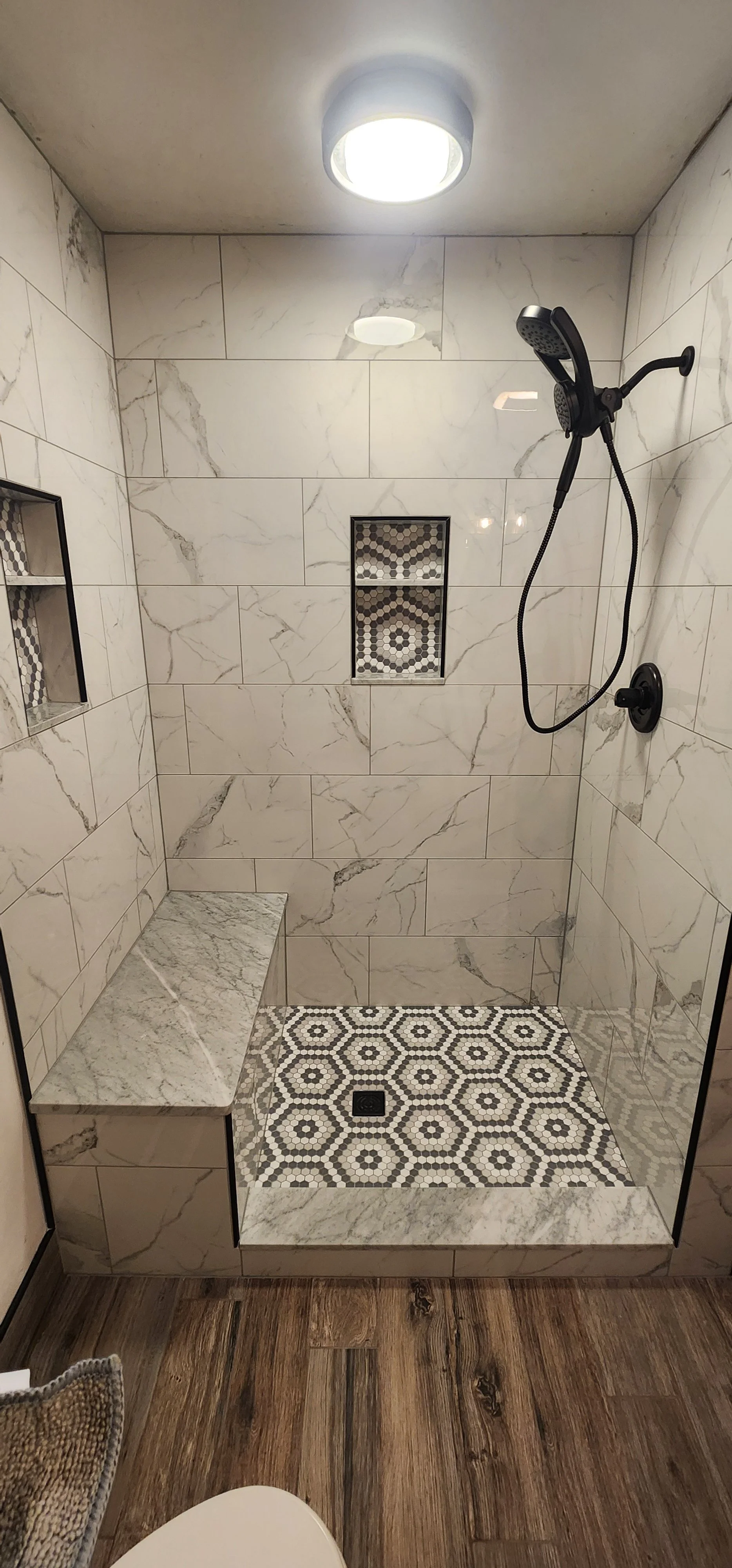 A modern walk-in shower with marble tile walls, a built-in marble bench, a patterned tile floor, a niche with marble and mosaic tiles, and a black showerhead mounted on the wall.