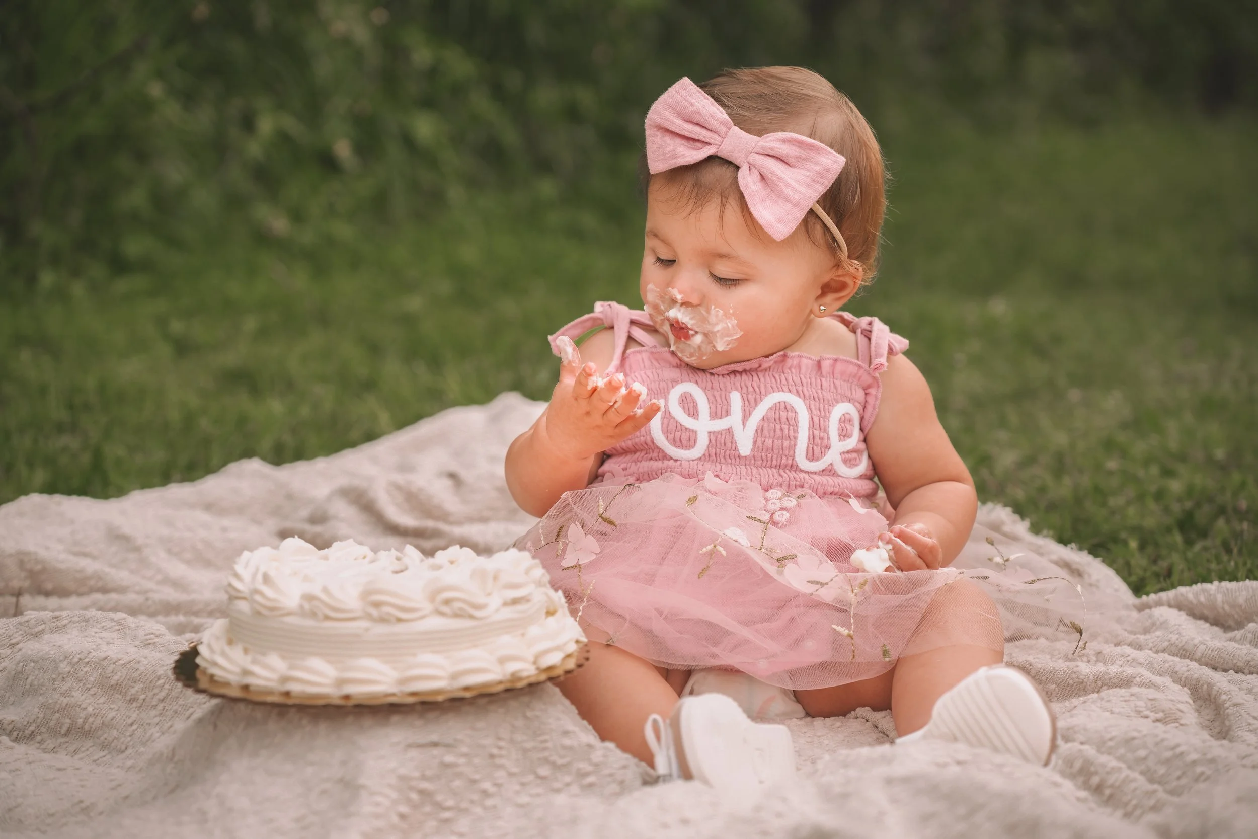 A young girl with light brown hair wearing a pink dress with the word 'Love' on it, sitting on a blanket outdoors. She has a pink bow headband and is eating a piece of cake, which has been smeared on her face and hands. In front of her is a white frosted birthday cake with decorative piping.