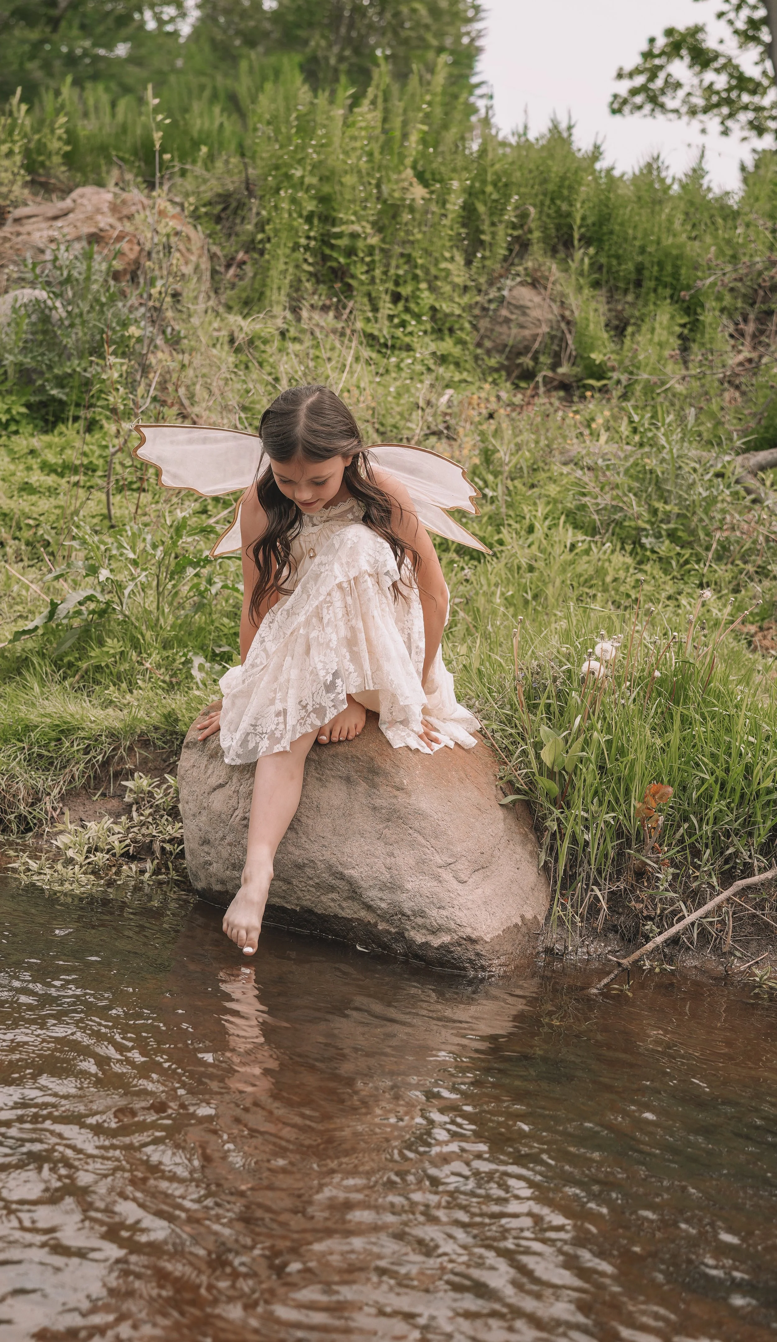 A young girl dressed as a fairy with wings, wearing a lace dress, sitting on a large rock at the edge of a creek, dipping her bare foot into the water. The lush green surroundings include bushes and trees.