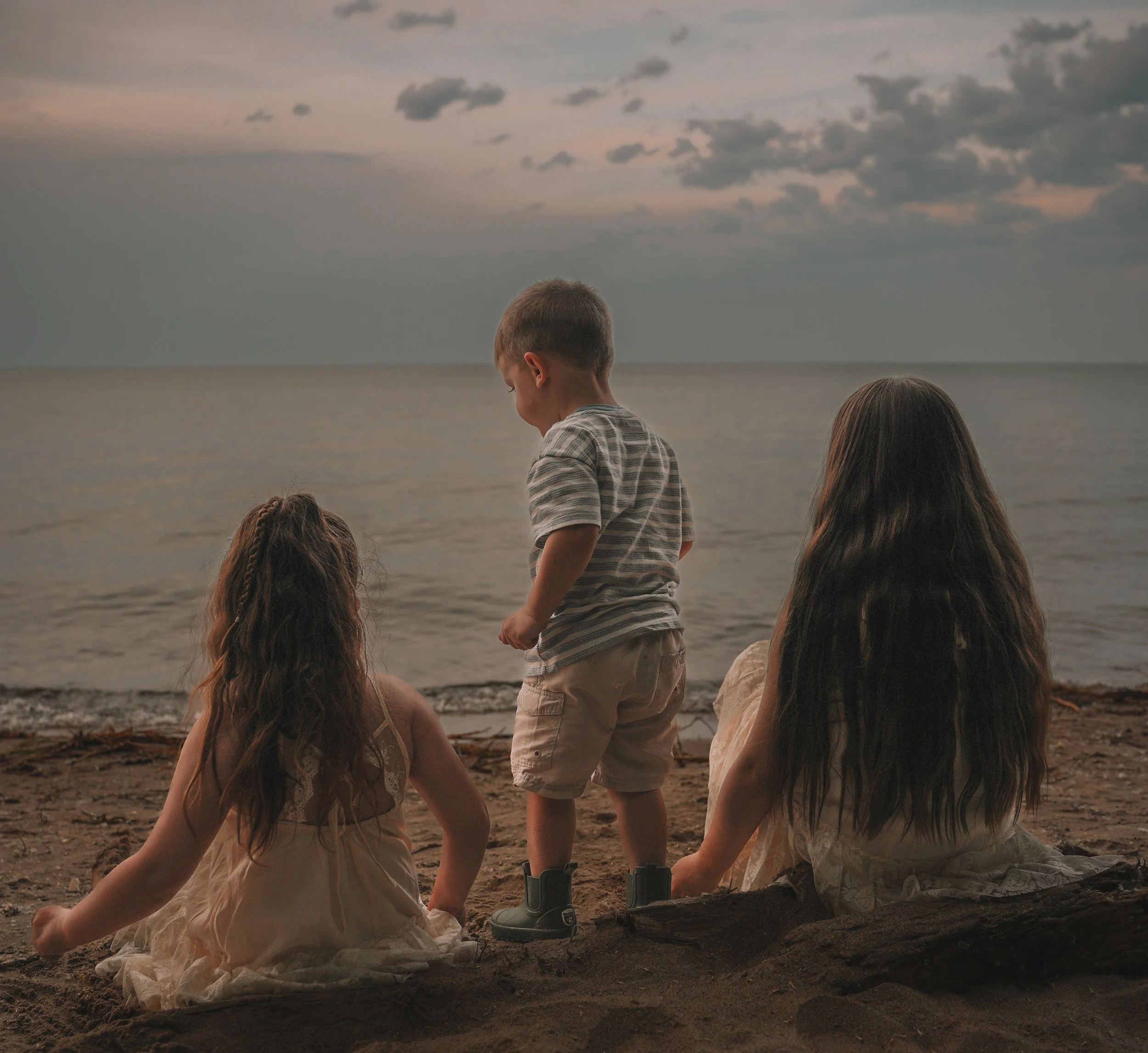 Two children by the beach at sunset, one sitting on the sand and the other standing, looking down, with the ocean and cloudy sky in the background.