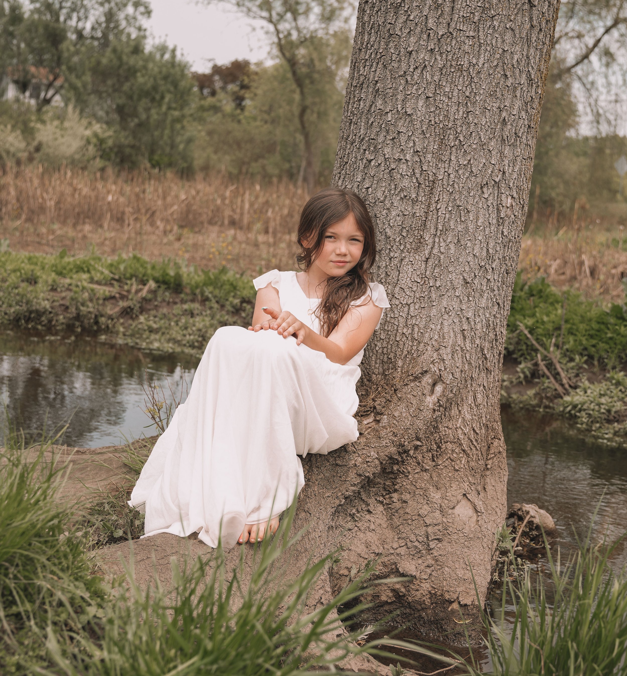 A young girl with brown hair wearing a white dress sitting by a large tree near a river, with greenery and trees in the background.