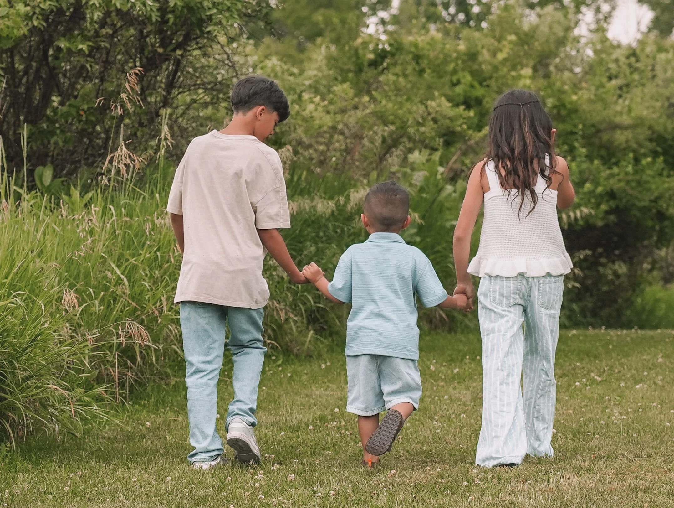 Three children holding hands walking in a grassy field with trees in the background.