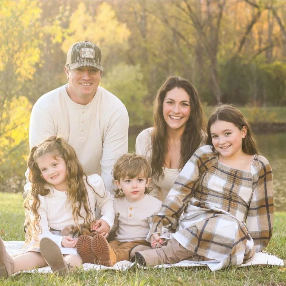 A happy family of five sitting on the grass outdoors in fall, near a lake, smiling at the camera. The father wears a cap and a cream sweater. The mother has long brown hair and is dressed in white. The three children, two girls and a boy, are dressed in fall clothing and are sitting on a white blanket.
