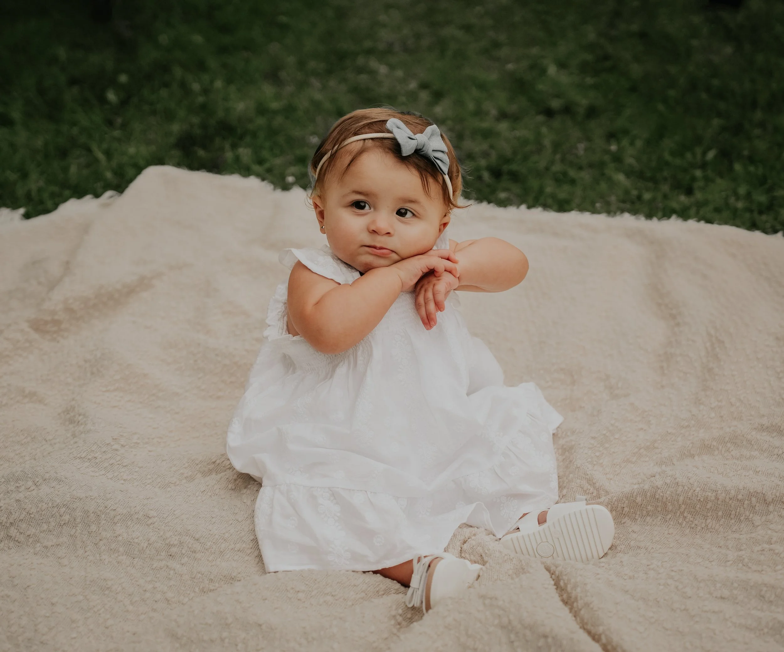 A young girl with light brown hair, wearing a white dress with lace details, white sneakers, and a gray bow headband, is kneeling on a beige carpet in front of a green grassy background, resting her chin on her hands and looking slightly to the side.