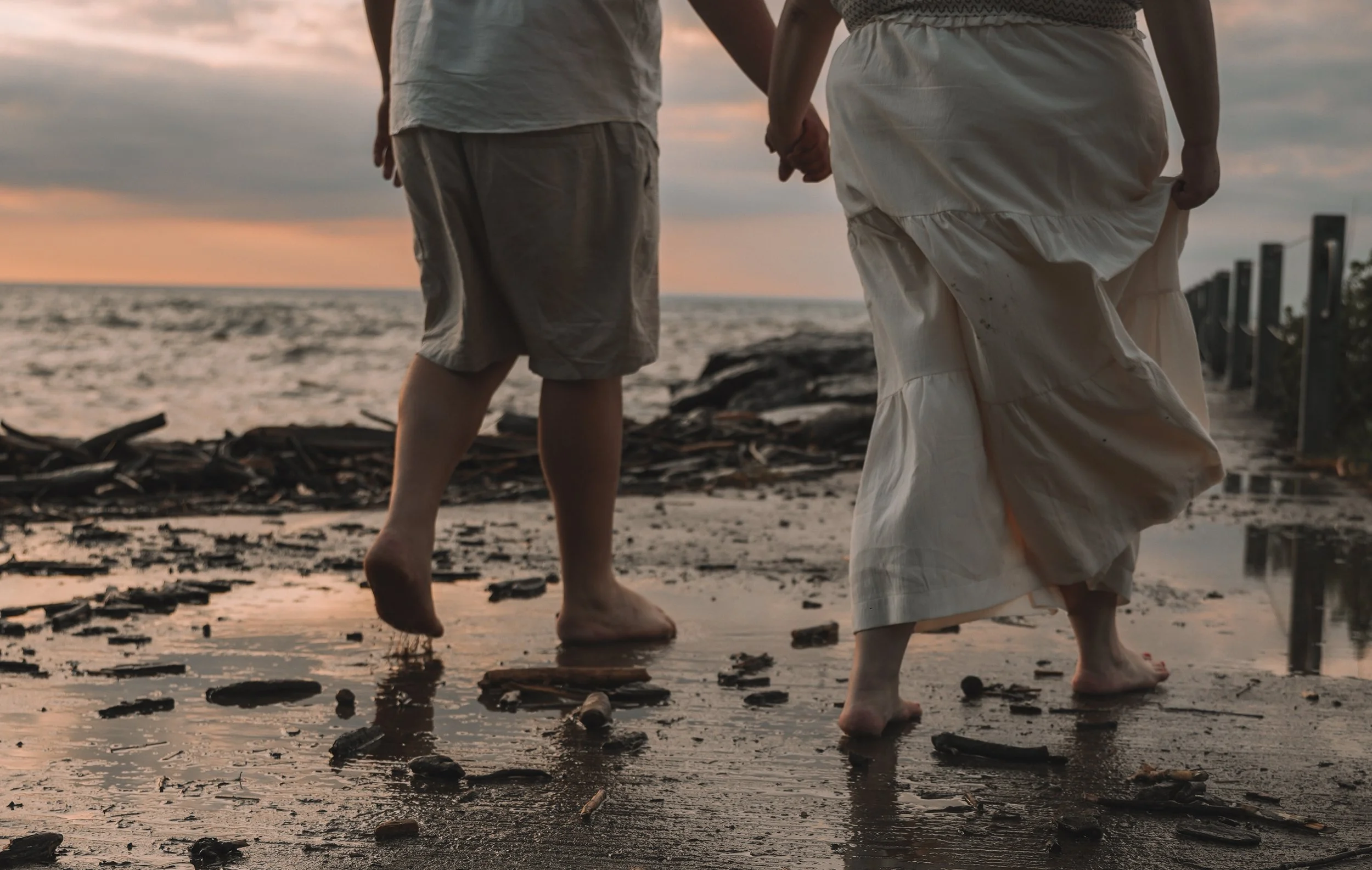 A couple walking hand-in-hand along a beach at sunset, with their feet in the wet sand and small rocks scattered around.