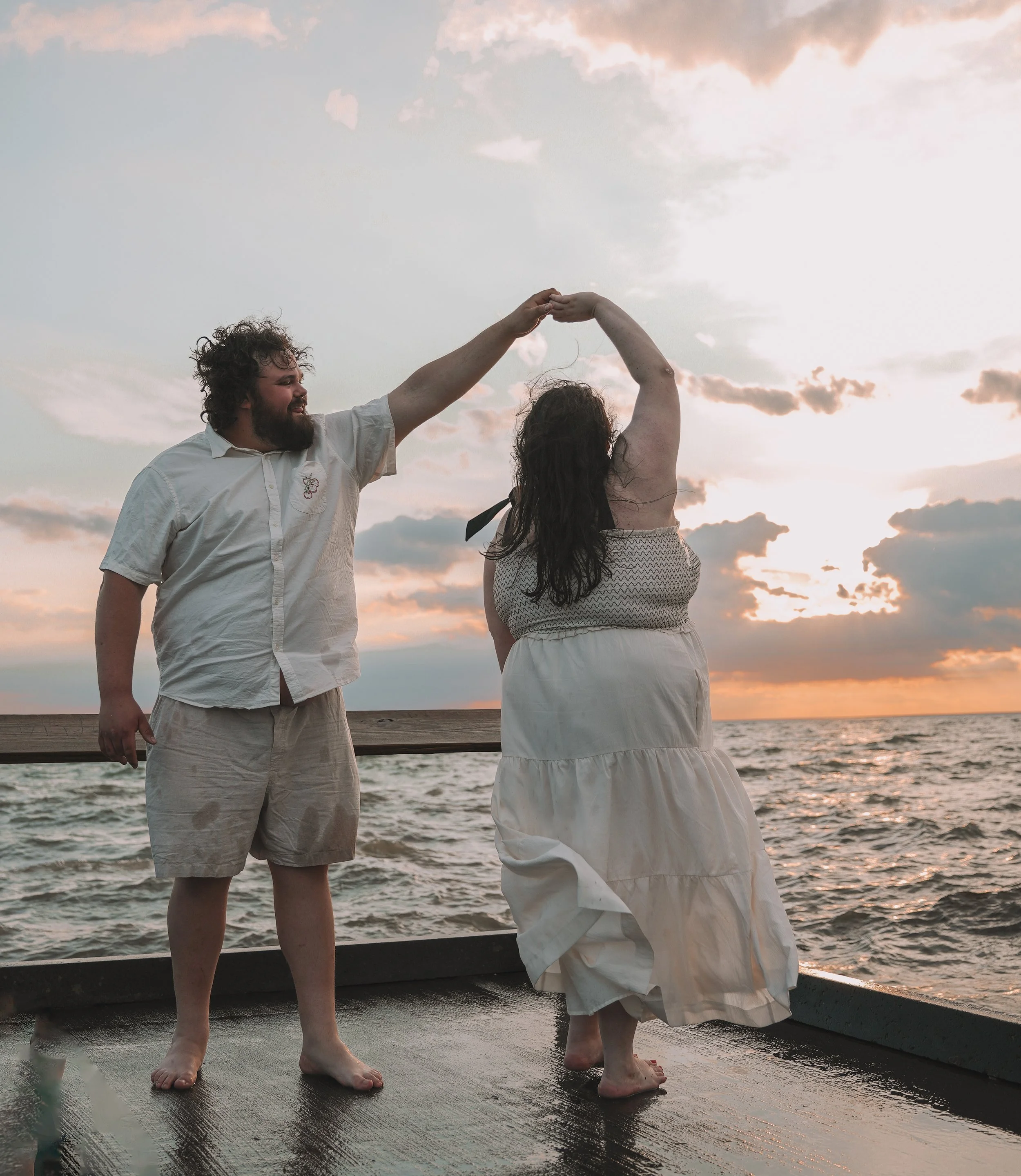 A couple dancing on a pier by the water at sunset, holding hands and smiling, with the sky filled with clouds and warm colors.