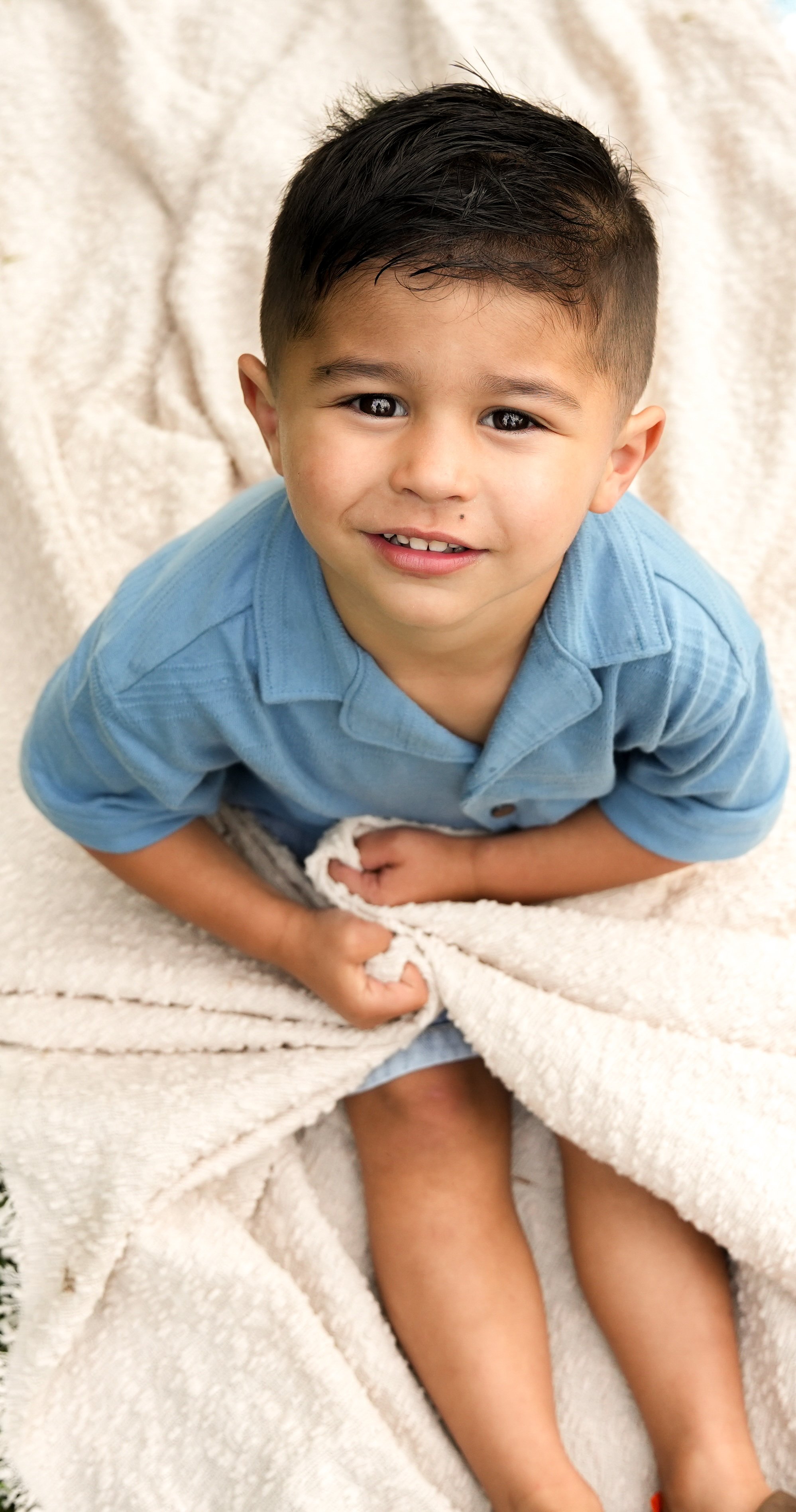 A young boy with dark hair and a light brown complexion sitting on a cream-colored blanket, wearing a blue shirt, holding the blanket with both hands, smiling at the camera.