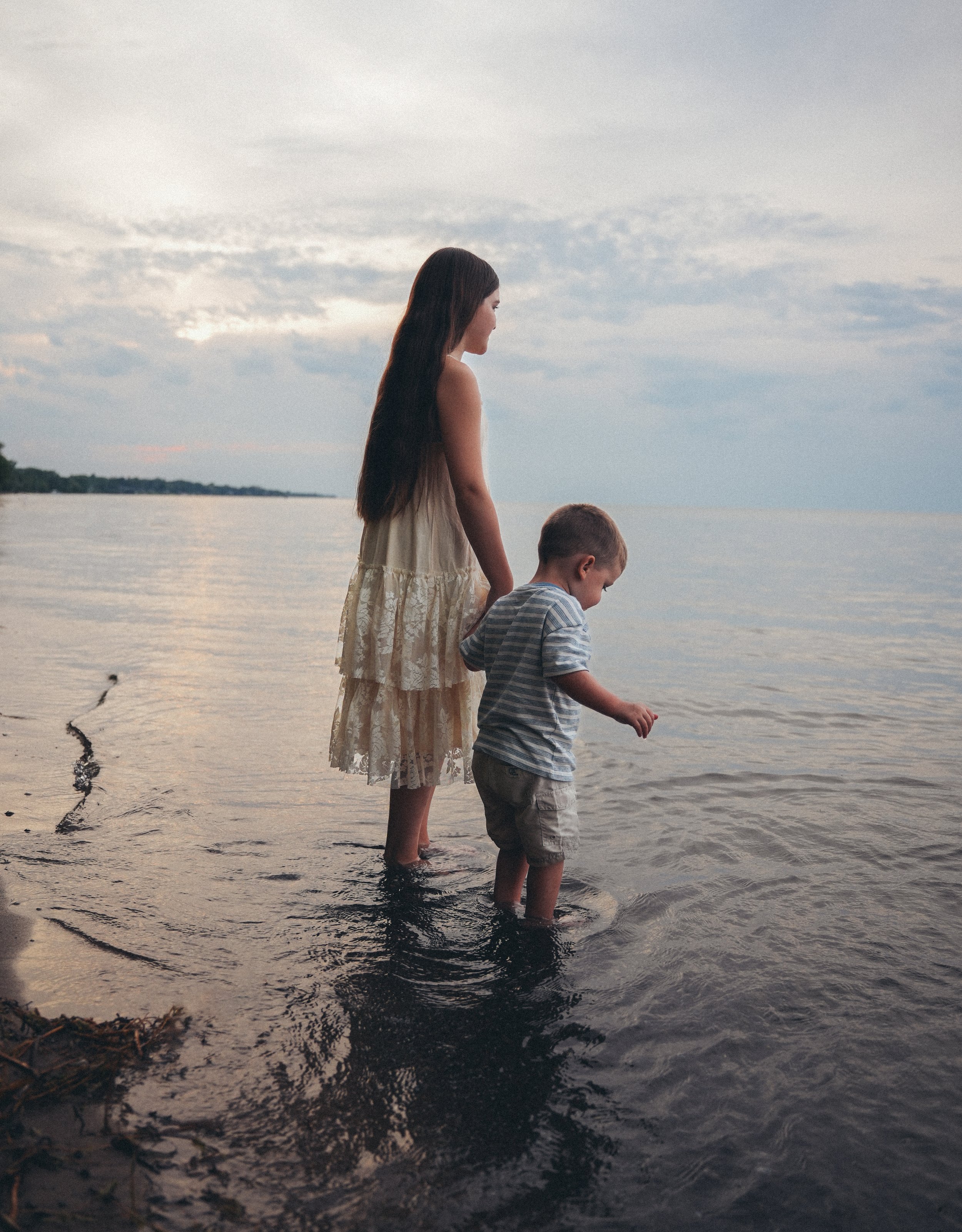 A sister and a younger brother standing in shallow water at the beach during sunset.