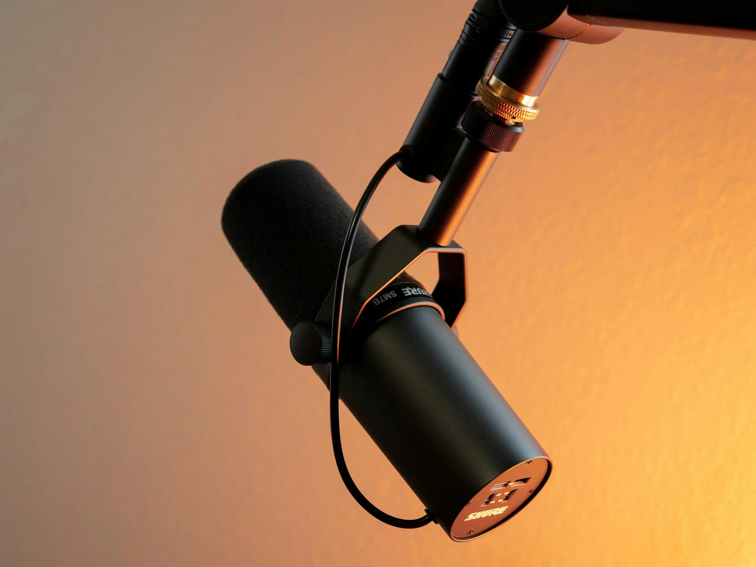 Close-up of a professional microphone mounted on an adjustable arm, with a foam windscreen and a black cable, against a gradient orange background.