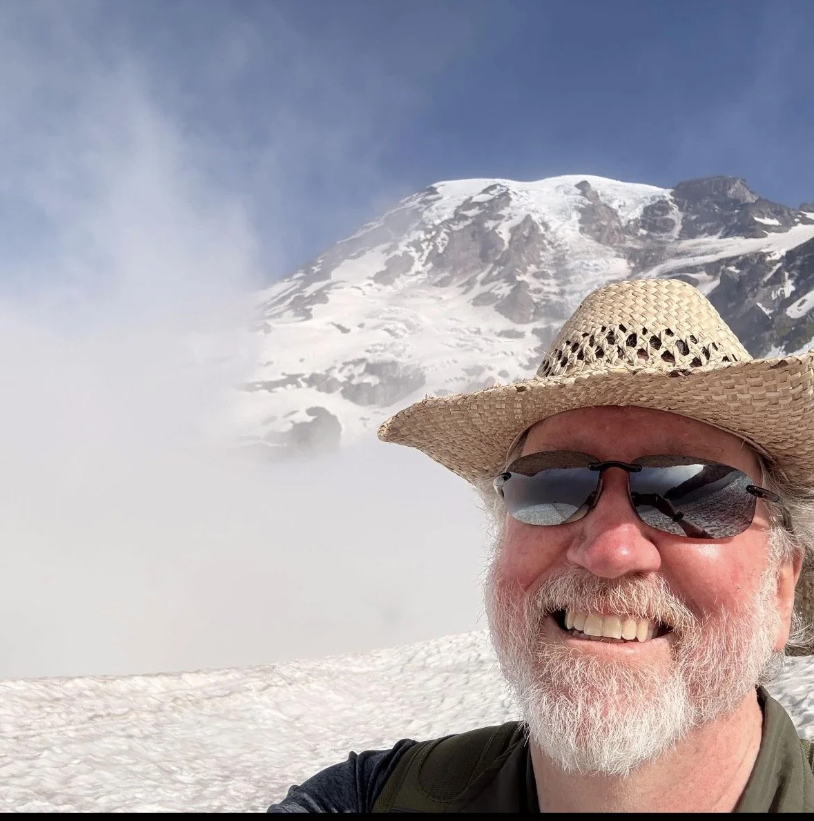 A smiling man with a beard, wearing sunglasses and a straw hat, takes a selfie with a snowy mountain in the background.