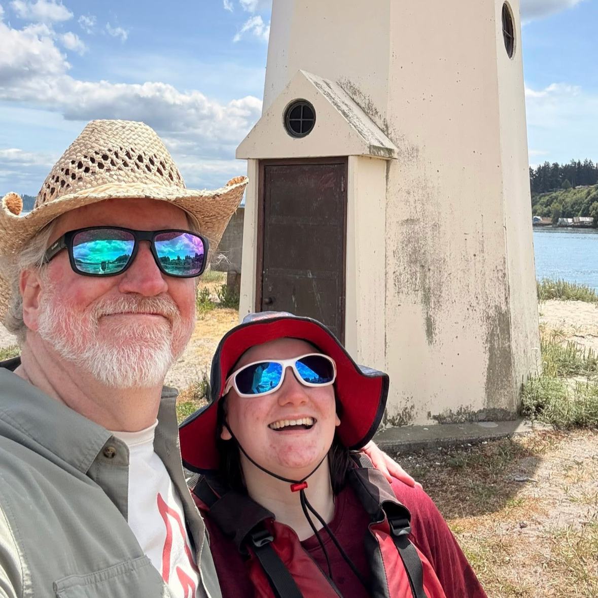 A man and a woman taking a selfie near a small lighthouse by the water. The man is wearing sunglasses, a straw hat, and a light-colored jacket; the woman is wearing sunglasses, a red hat, and a red jacket. Both are smiling, standing outdoors on a partly cloudy day.
