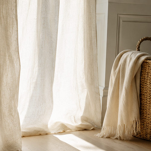 Sunlight filters through cream-colored curtains onto a hardwood floor, with a woven basket and a beige throw blanket nearby.
