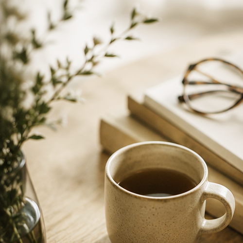 A cup of coffee on a wooden surface with a stack of books and reading glasses, next to a vase with greenery.
