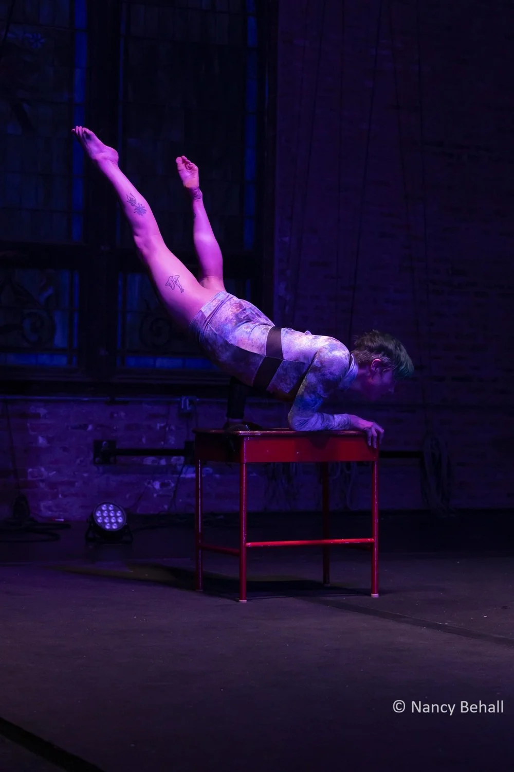 A circus performer with short green hair performs an arm balance on a small red table, balancing with their legs extended upward in a dimly lit setting with purple and blue lighting. They wear a sparkly long sleeve leotard.