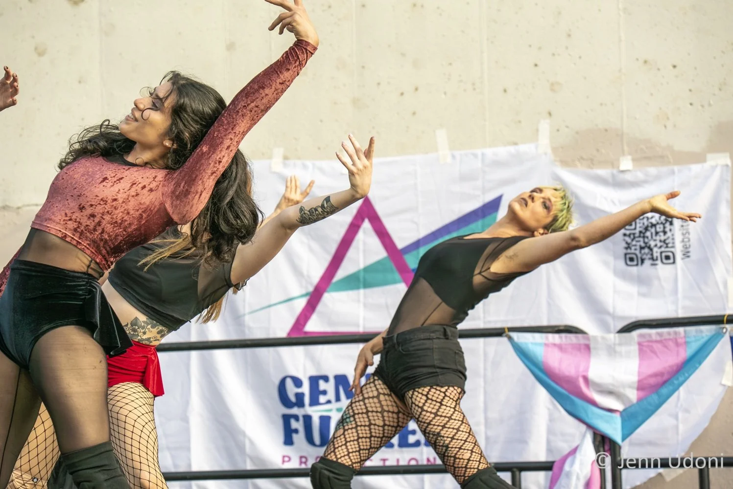 Three dancers perform on stage with their arms raised and their backs arched, wearing mesh shirts, fishnet stockings and black shorts, in front of a colorful banner with a trans pride logo and a QR code.