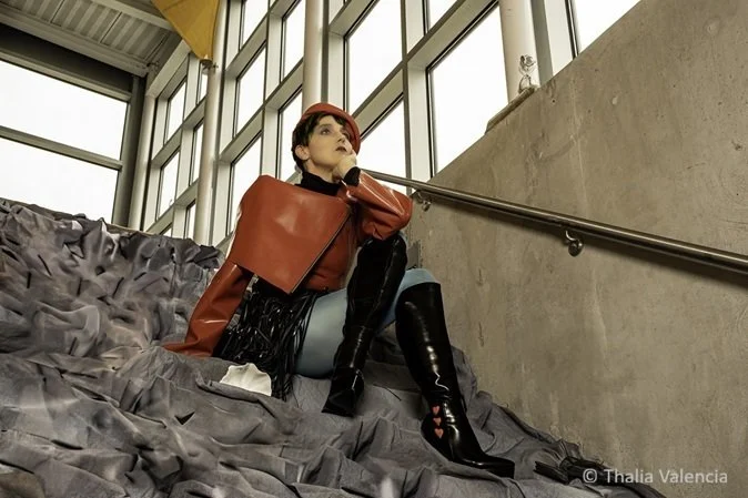 Fashion model with short brown hair sitting on a crumpled gray fabric surface inside a modern building with glass windows, wearing a red jacket, matching red hat, black knee-high boots with heart details.  
