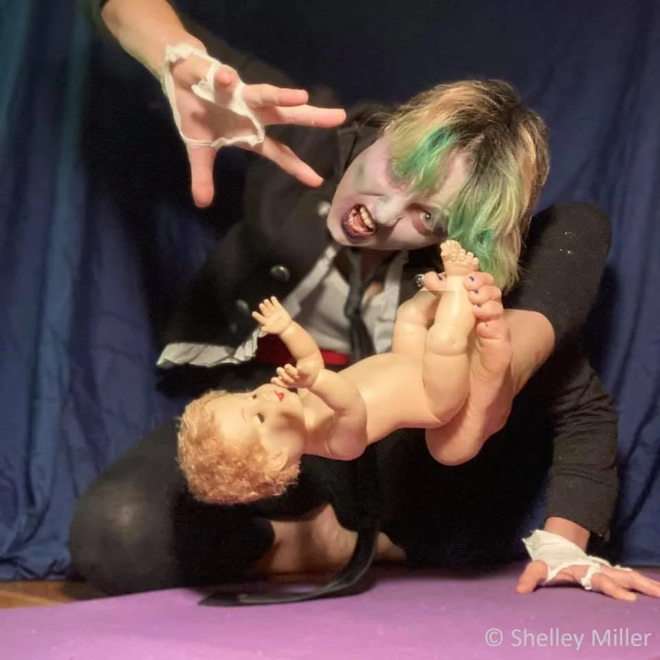 Person with green dyed hair making a playful face, holding a realistic-looking doll with curly blonde hair and a pacifier. The person is wearing a black jacket and has bandages on their fingers. They are on a purple mat with a blue curtain or backdro
