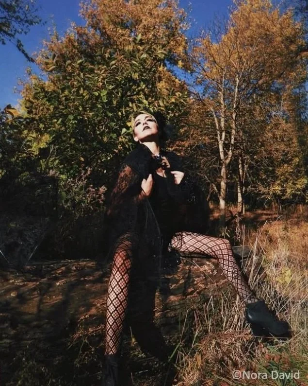A person with makeup and dark clothing sitting outdoors on a rock, with trees and a blue sky in the background.