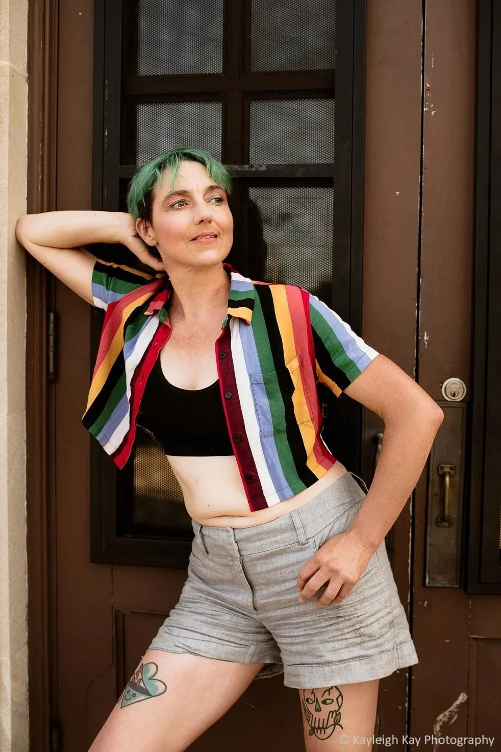A person with short green hair posing in front of a door, wearing a rainbow-striped short-sleeve shirt, black crop top, and gray shorts, with tattoos on their thighs.