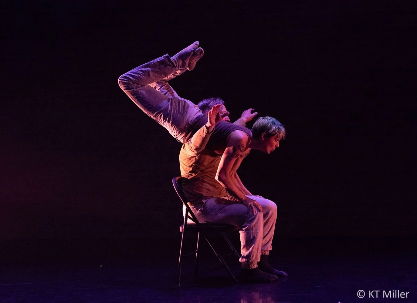 Two performers are on a dark stage, one sitting on a chair and the other balancing on their back, forming a human structure in an artistic pose.